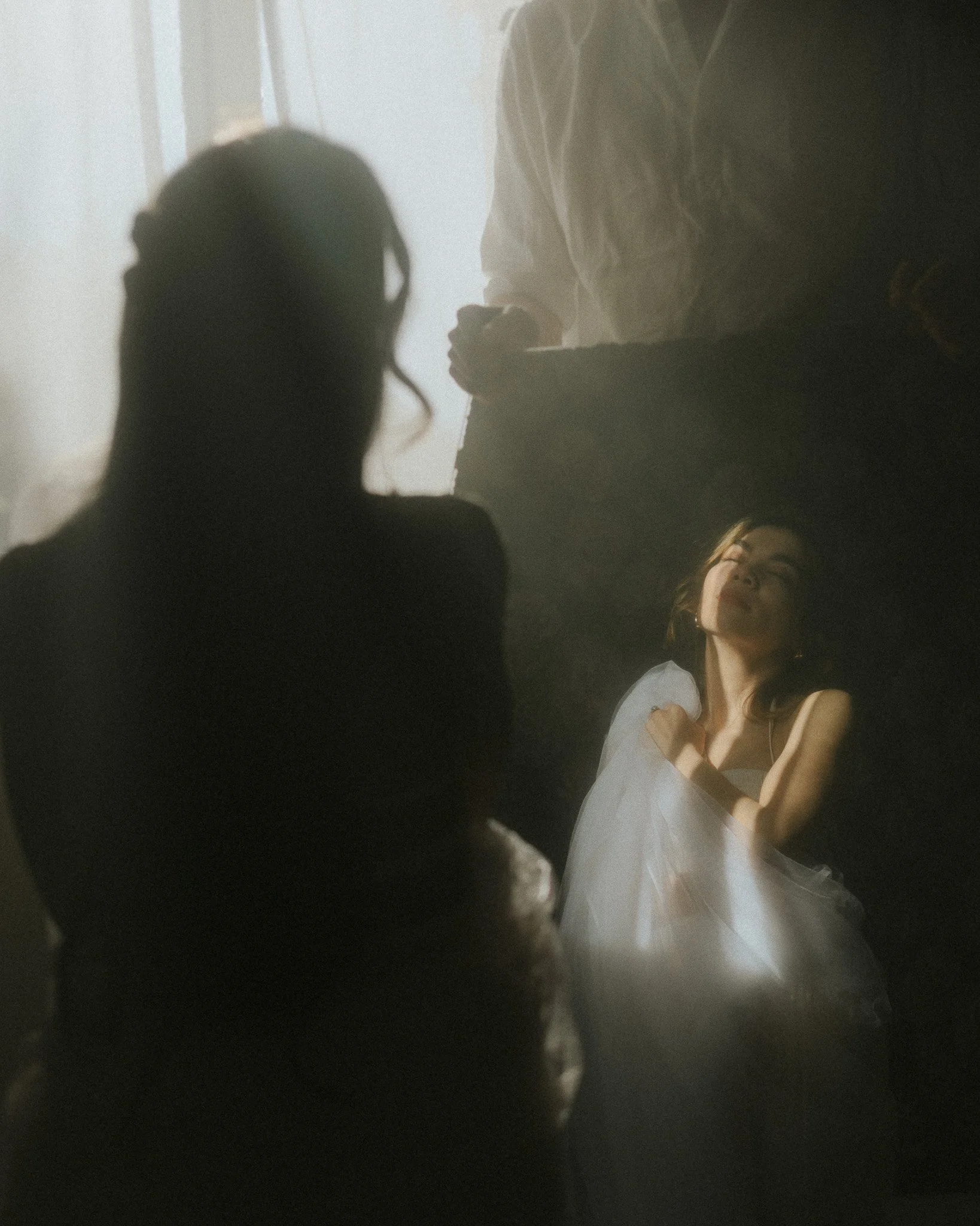 Eine Frau in einem weißen Kleid liegt auf dem Boden und blickt nach oben, während zwei Personen im Vordergrund unscharf sind, die auf sie schauen, mit Sonnenlicht, das durch ein Fenster scheint.