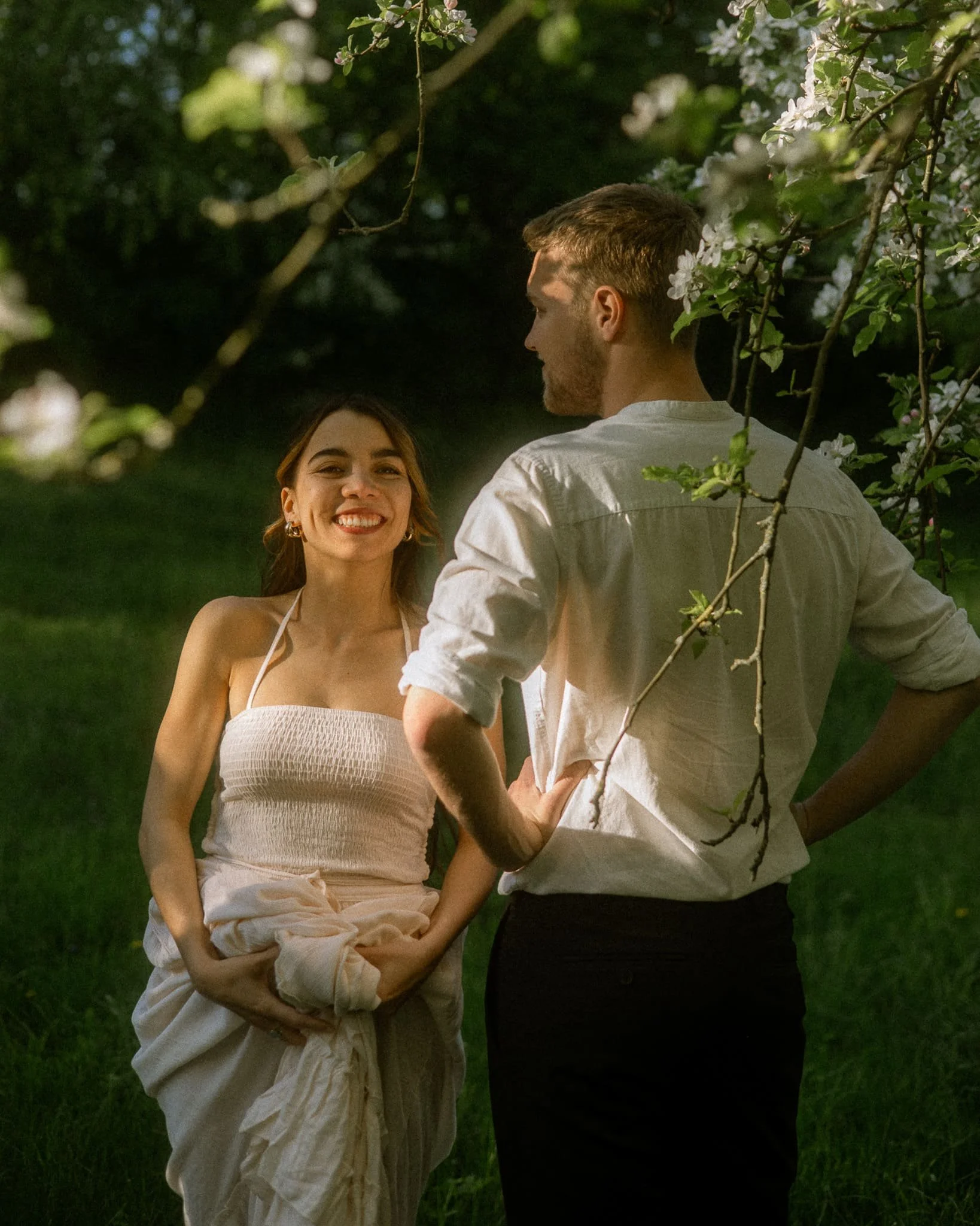 Ein lachendes Mädchen in einem weißen Sommerkleid steht im Grünen, während ein Mann mit weißen Hemd ihr gegenübersitzt und lächelt, umgeben von blühenden Zweigen.