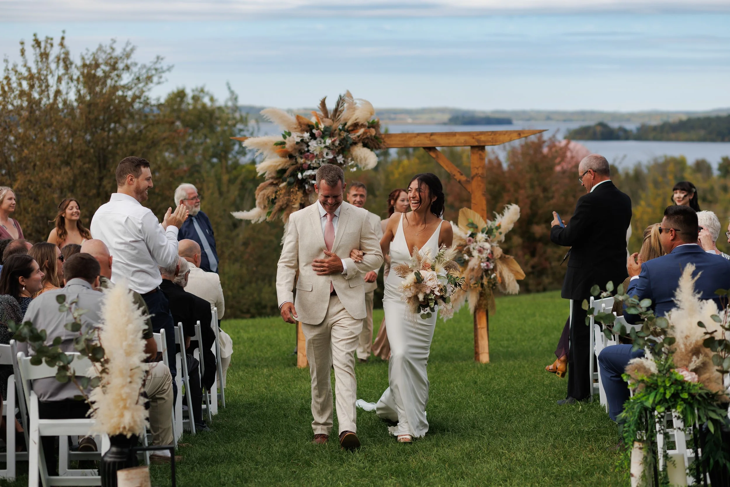 A wedding ceremony outdoors by a lake with a forested backdrop. The bride and groom are walking down the aisle, smiling and holding hands. The bride is wearing a white gown and holding a bouquet, while the groom is in a light-colored suit. Guests are seated on either side, some clapping and taking photos.