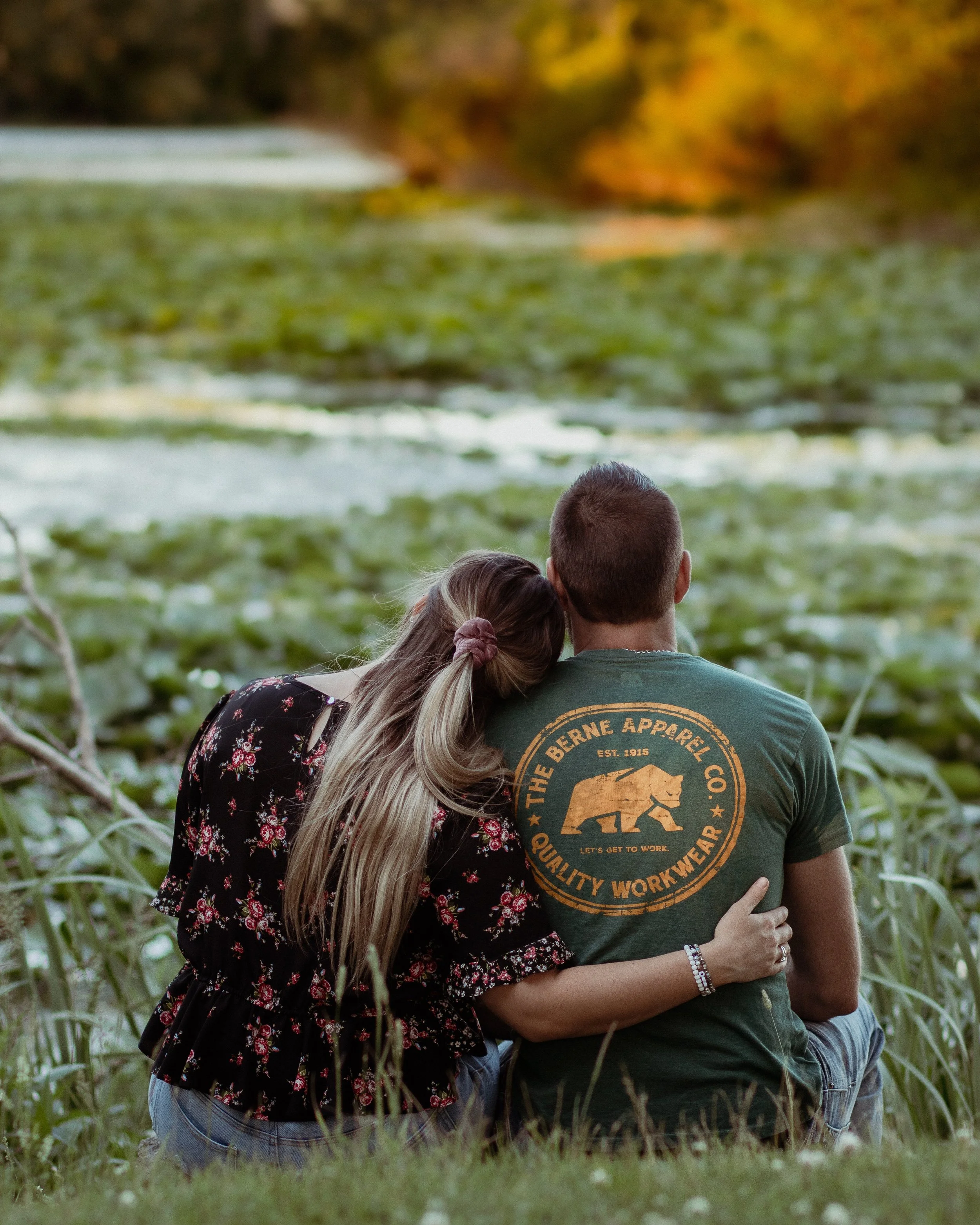 A couple sitting on grass near a pond during autumn, with the woman resting her head on the man's shoulder, both facing away from the camera.