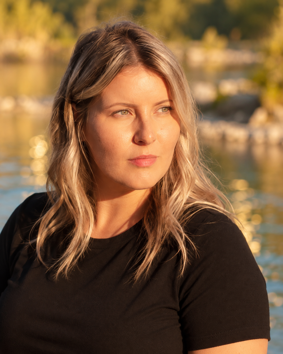 A woman with blonde wavy hair looking to the right outdoors near a body of water during golden hour.