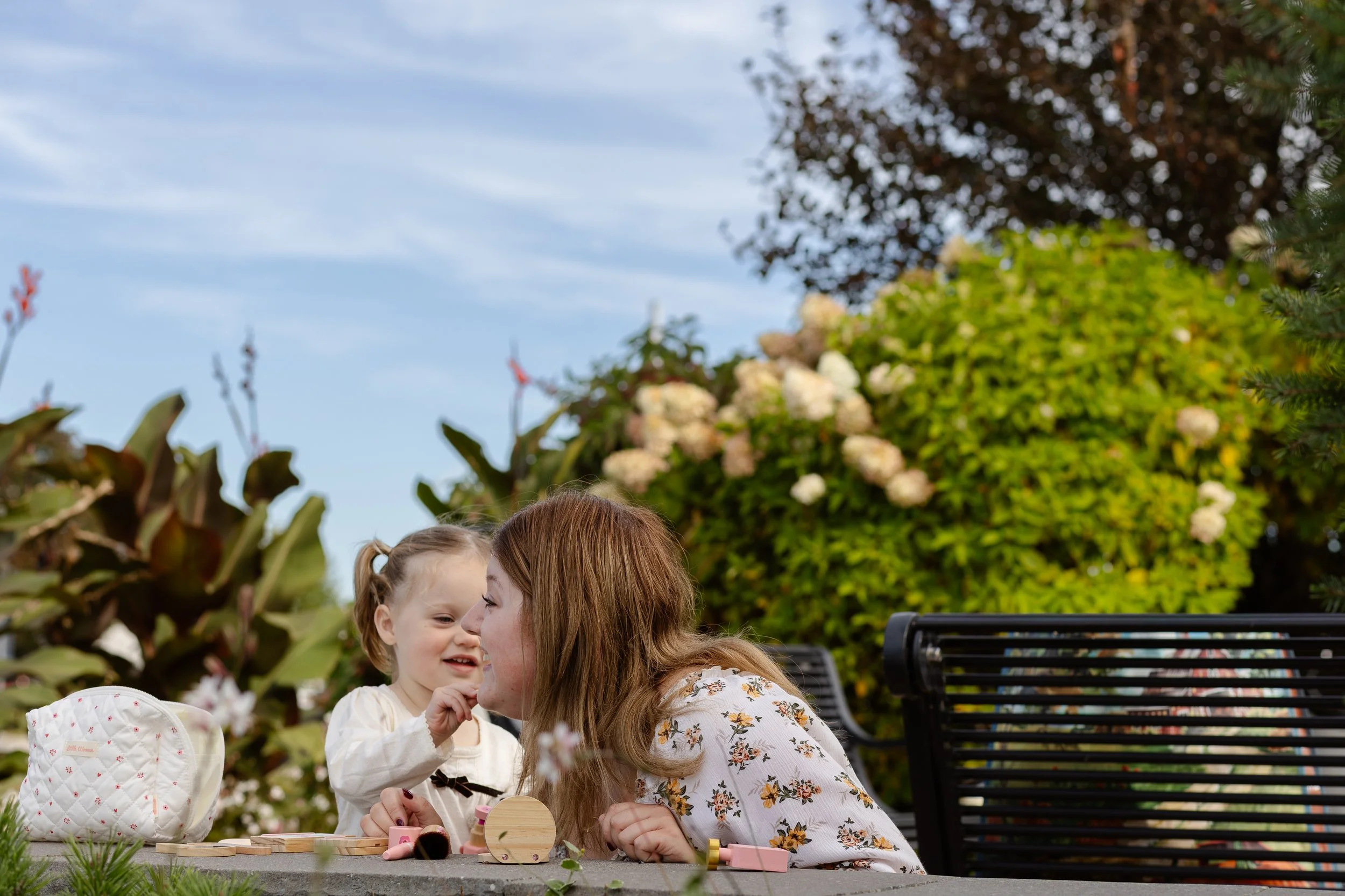 A woman and a young girl are sitting outdoors in a garden, smiling and playing with small toys. The garden is lush with green foliage and blooming white flowers, and there are trees and a bench visible in the background. The Tied Vows, documentary we