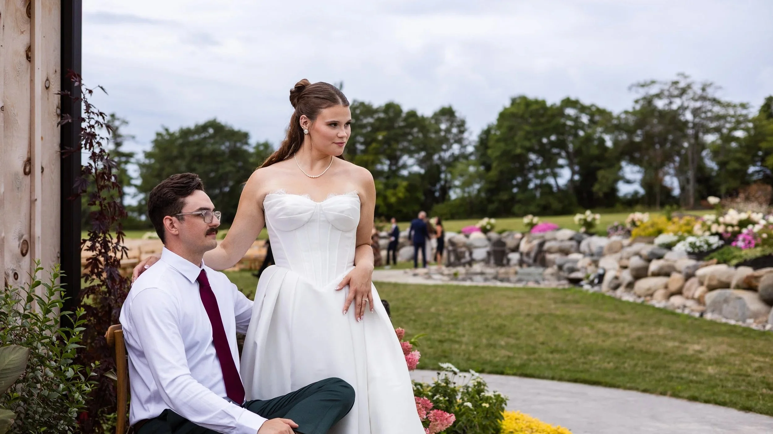 A bride standing beside a seated man at an outdoor wedding ceremony, with a grassy area, trees, and flowers in the background.