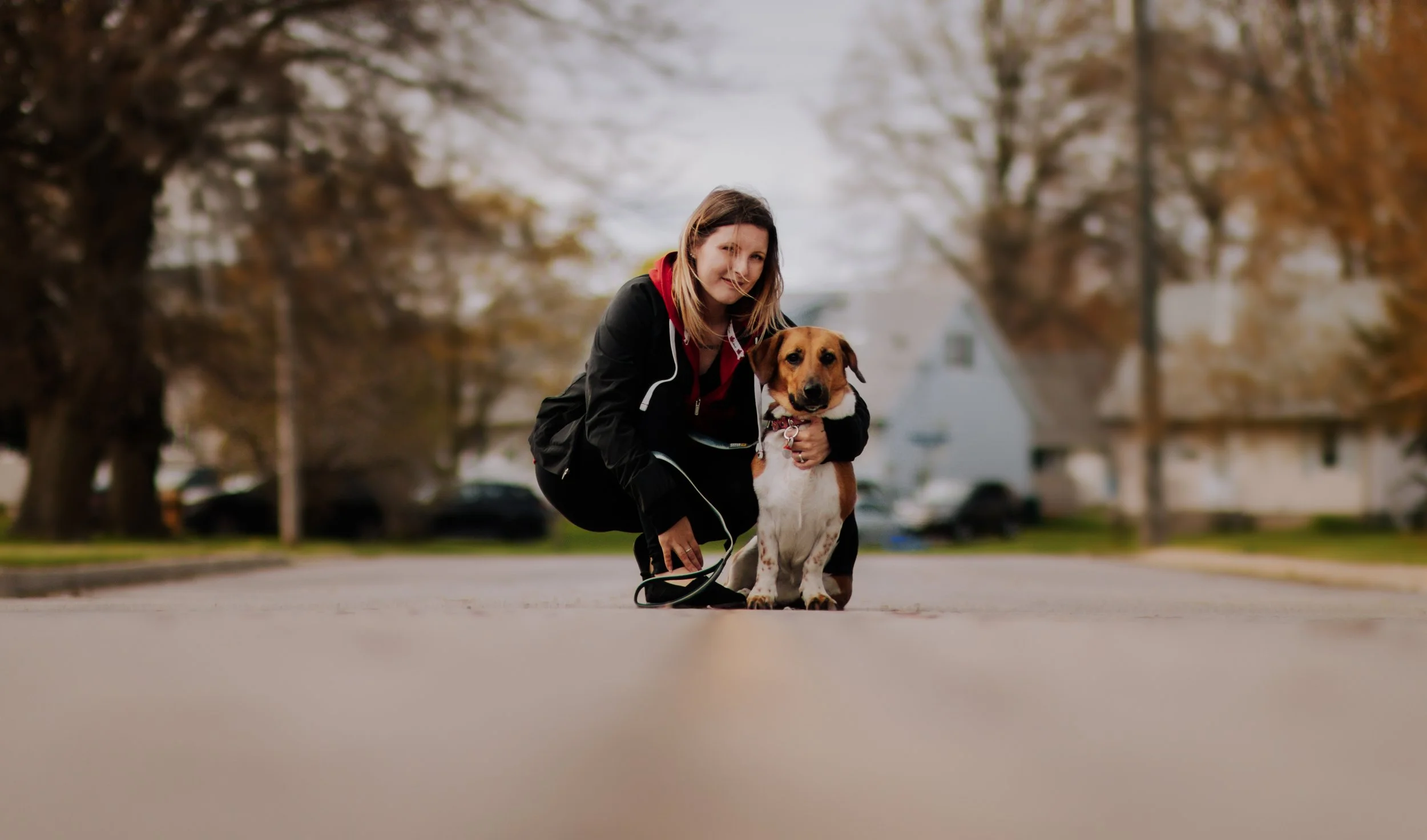 A woman squatting on a quiet street during fall, holding a brown and white dog, with trees and houses in the background.