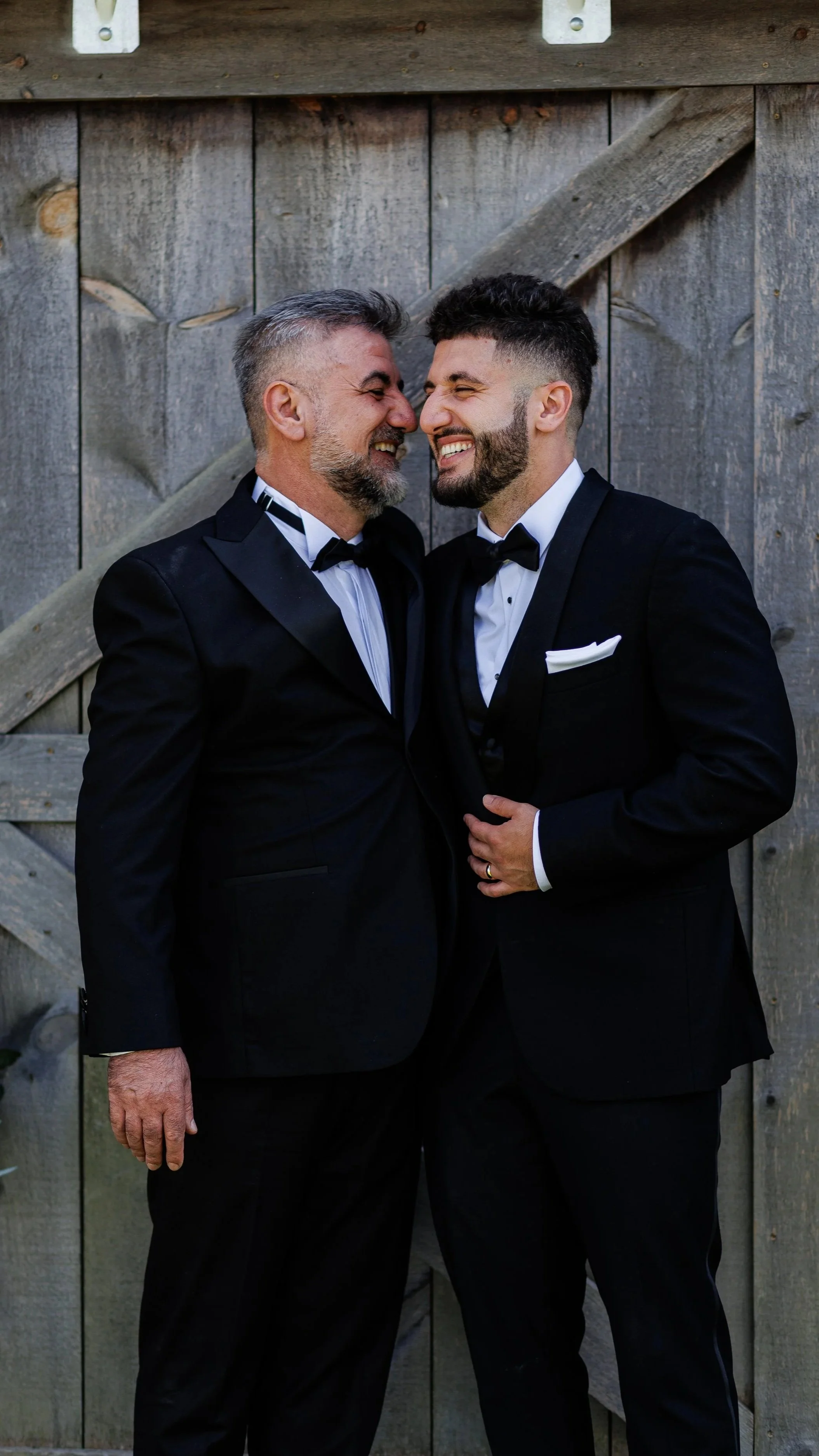 Two men in tuxedos embrace and smile, standing in front of a rustic wooden fence.