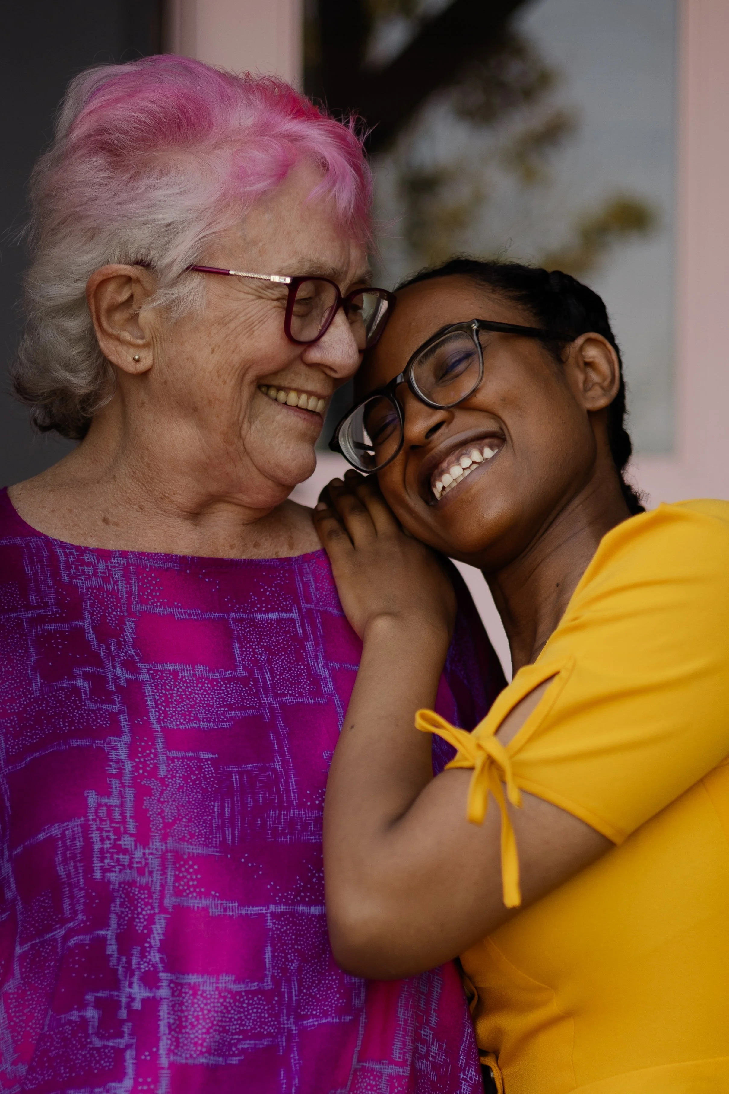 Two women sharing a joyful embrace, smiling with eyes closed, one older with white hair and glasses, the other younger with dark hair, glasses, and wearing a yellow top.