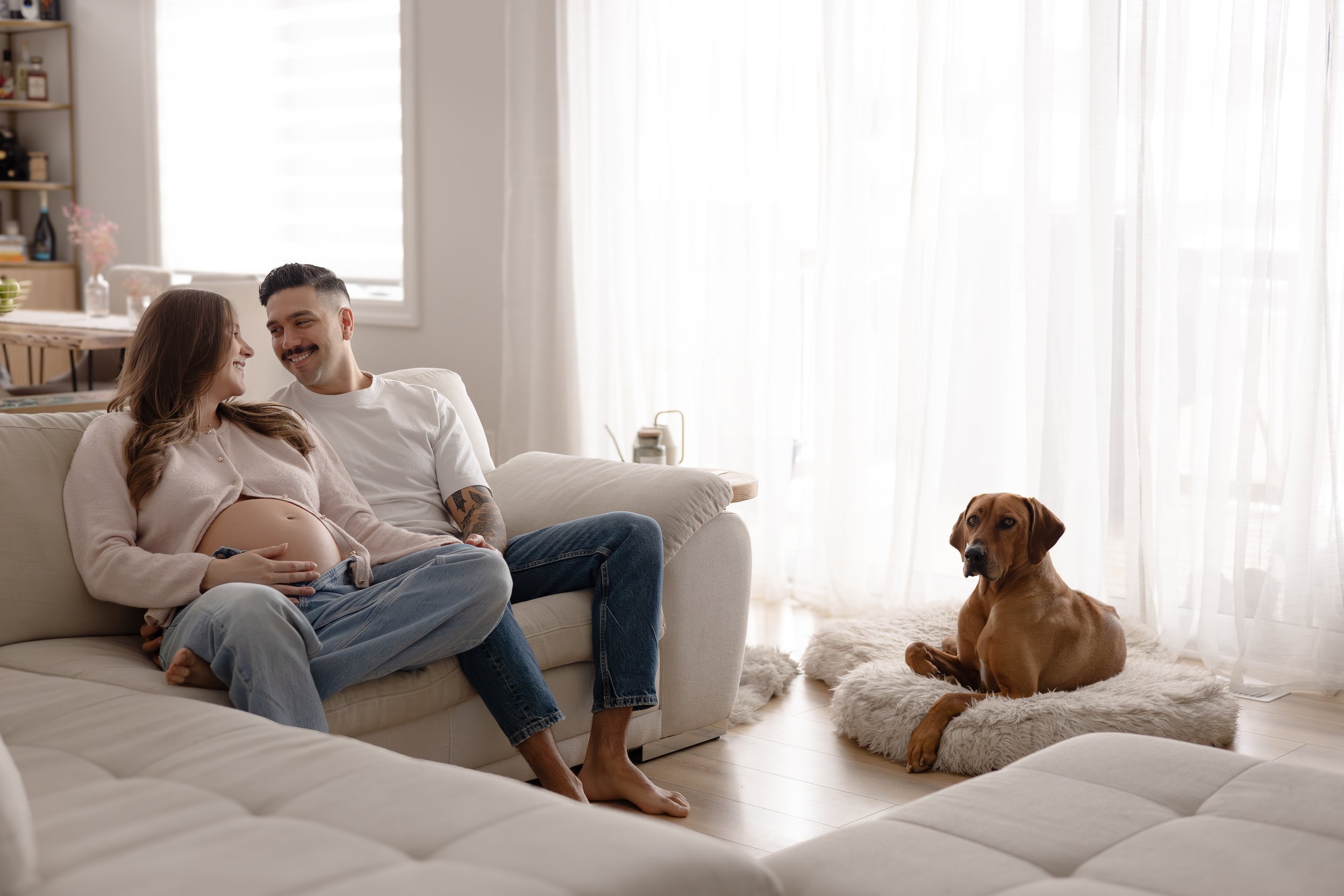 Pregnant woman and man sitting on a sofa while smiling at each other, dog lying on a fluffy mat nearby in a well-lit living room.