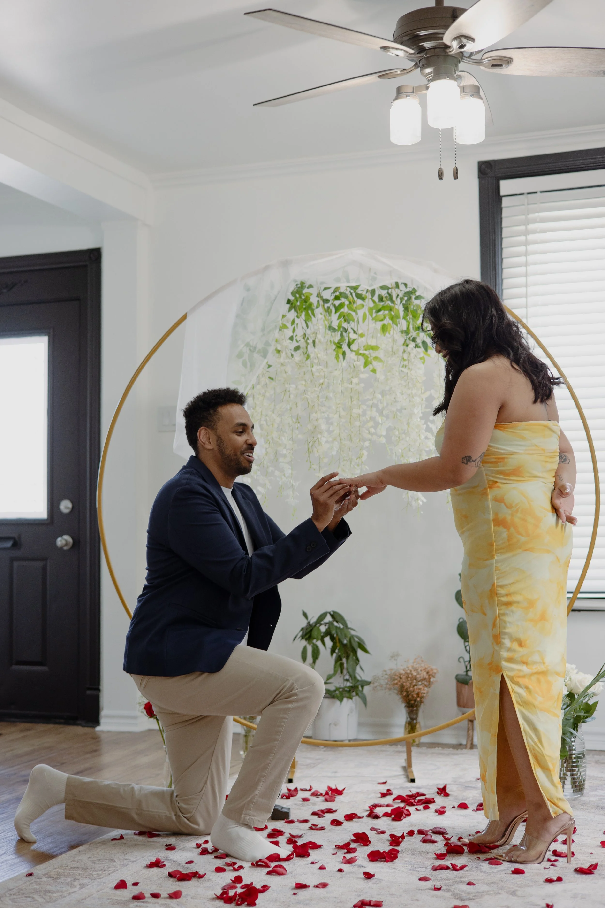 A man kneeling on one knee proposes to a woman inside a decorated room with rose petals on the floor.