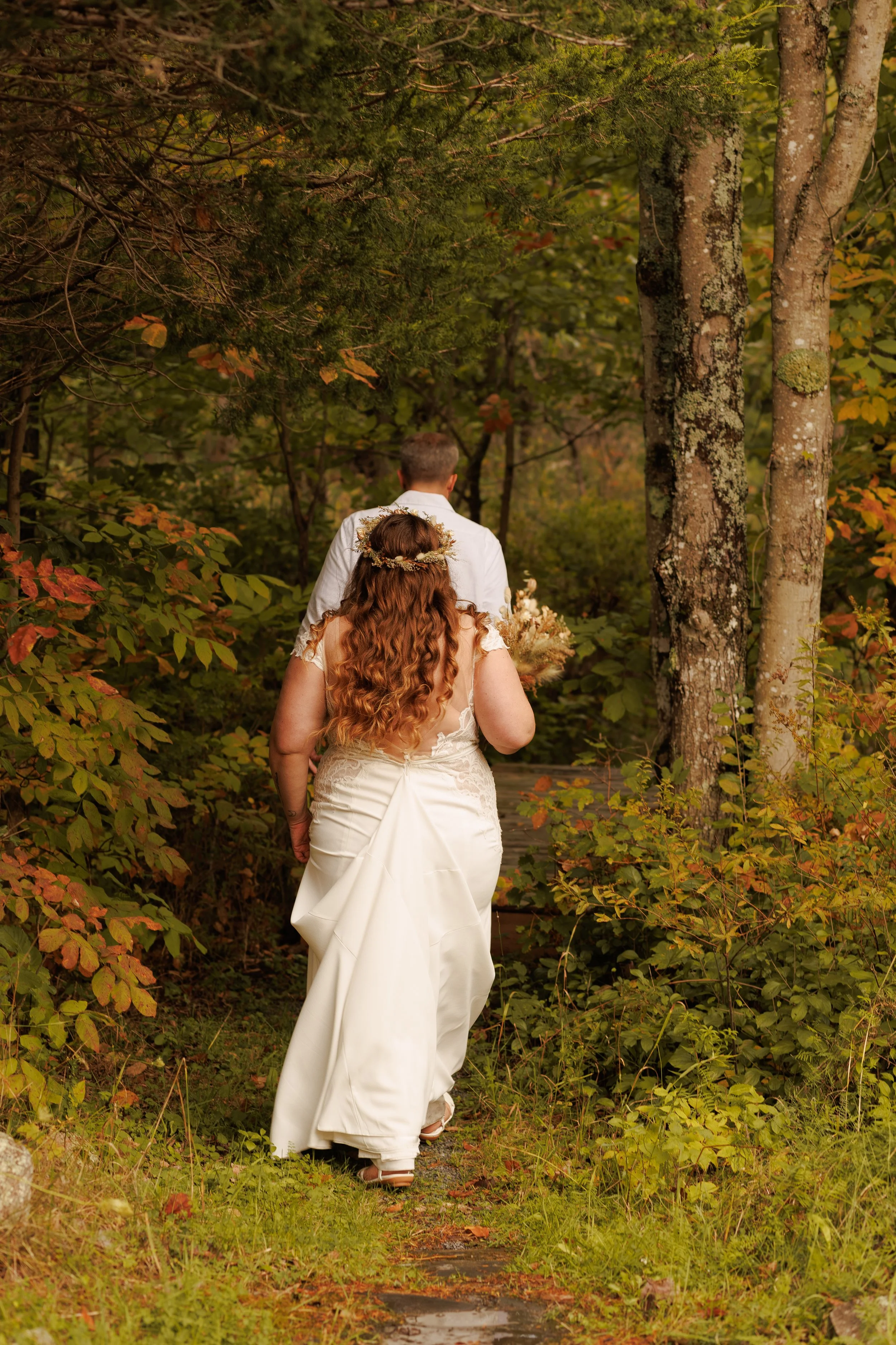 A bride and groom walking along a forest path, surrounded by autumn foliage, with the bride holding a bouquet and wearing a white wedding dress.