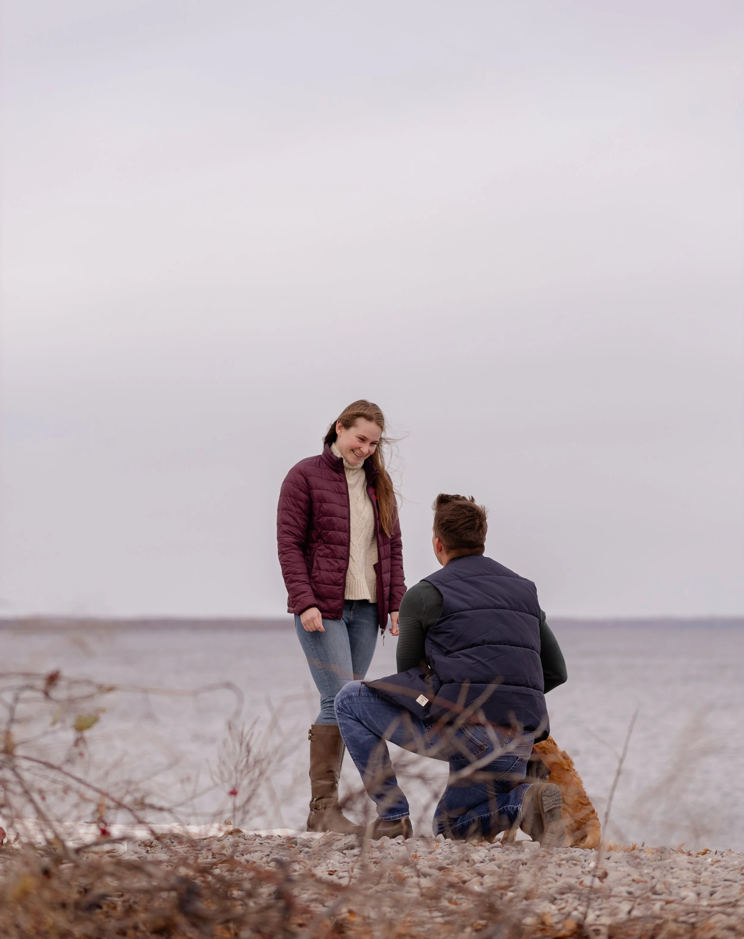A man proposing marriage to a woman on a rocky shoreline with water in the background. The woman is smiling, wearing a maroon jacket and jeans, the man is kneeling, wearing a dark vest and jeans, with a dog nearby.