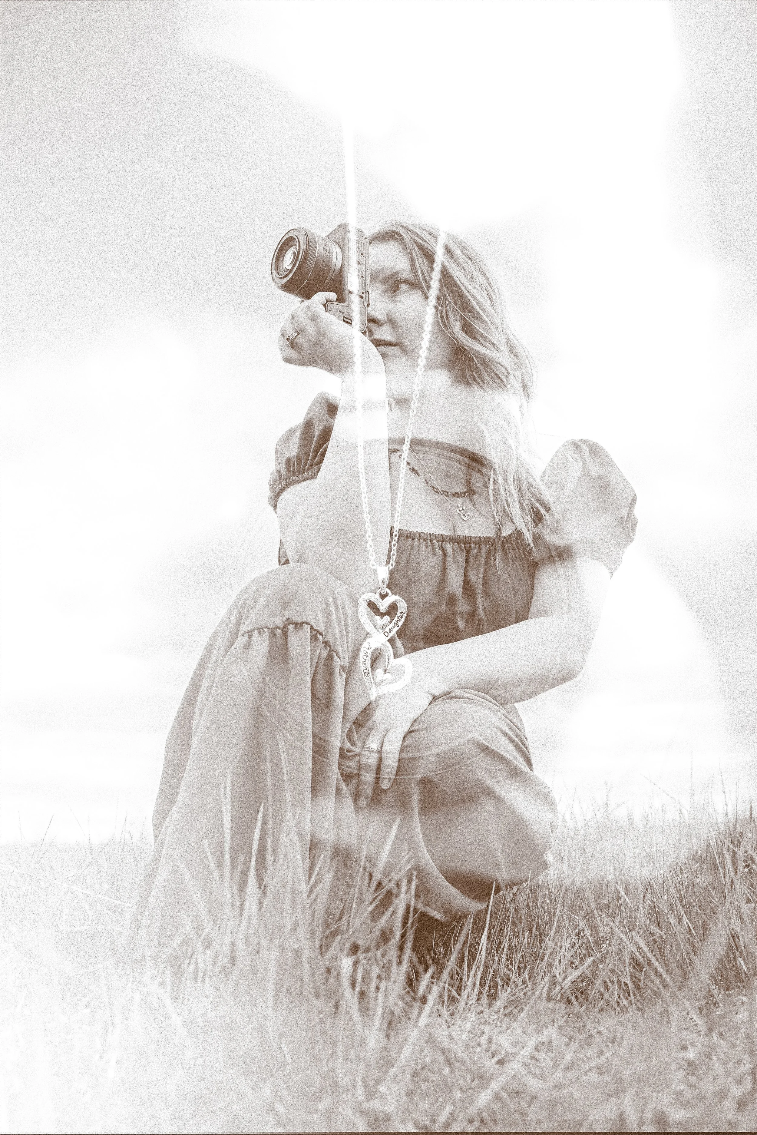 A young woman with wavy hair sitting on grass, holding a camera to her eye, with a necklace featuring two hearts hanging from her neck. The photo is in black and white with a high-key, bright background.