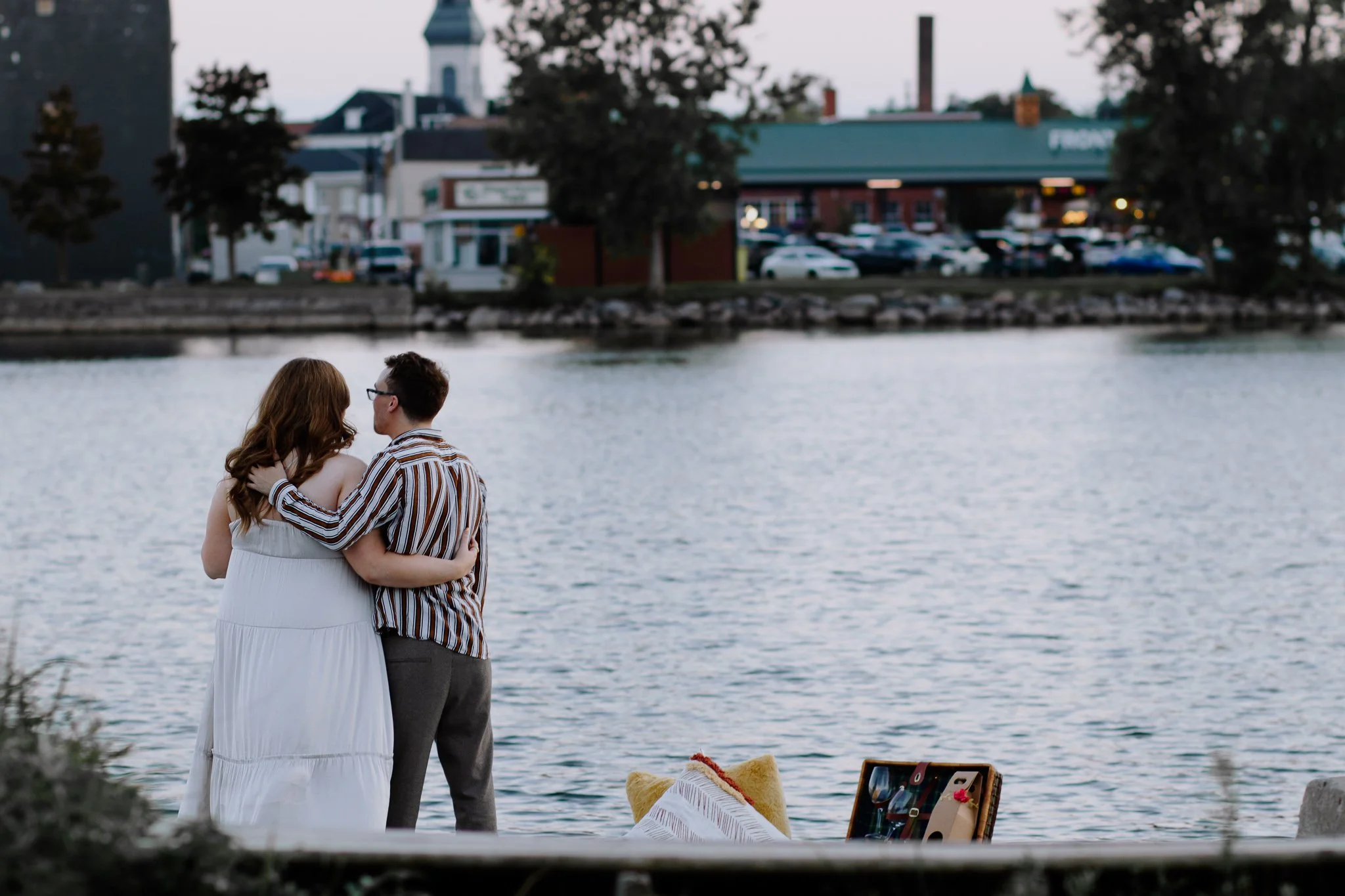 A couple standing by a river at sunset, embracing and looking at each other, with a small picnic setup nearby containing a cushion and a suitcase.