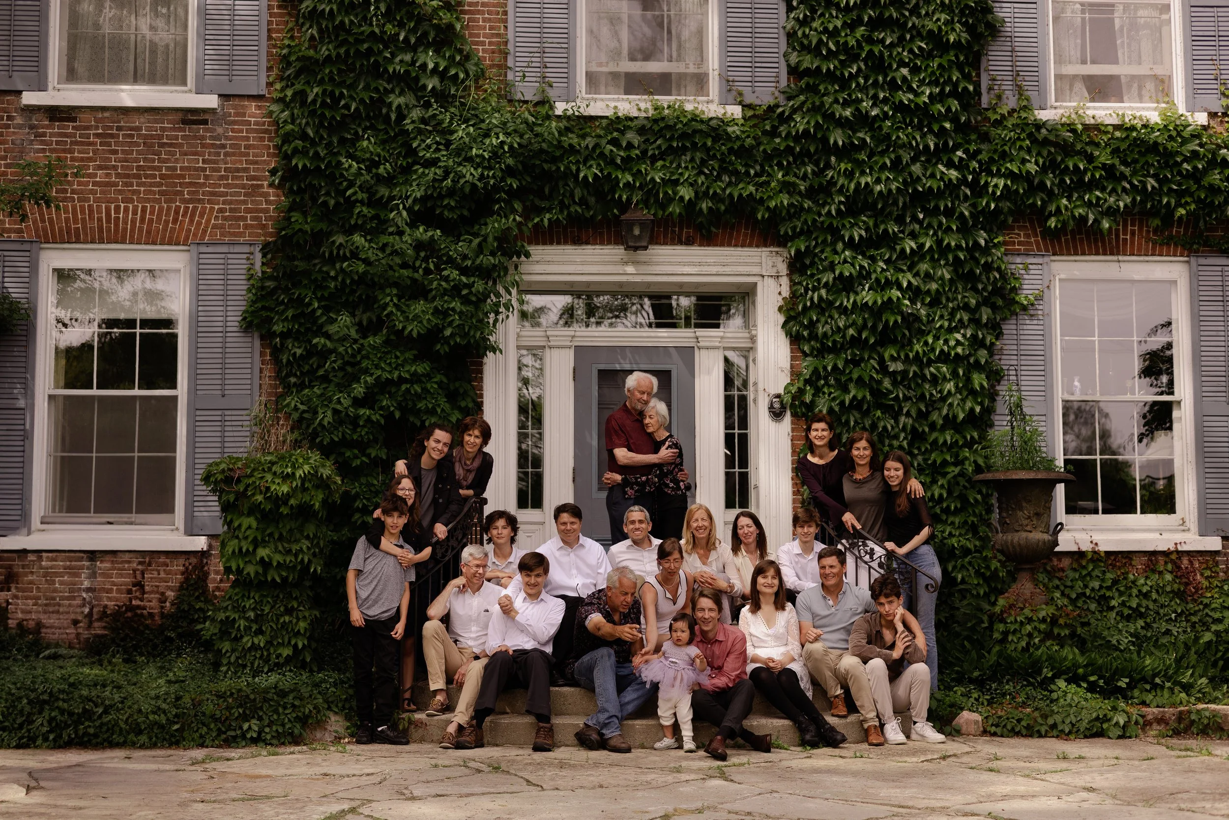 A large group of people, including children, adults, and seniors, gathered on the steps of a brick house covered in green ivy, posing for a group photo.