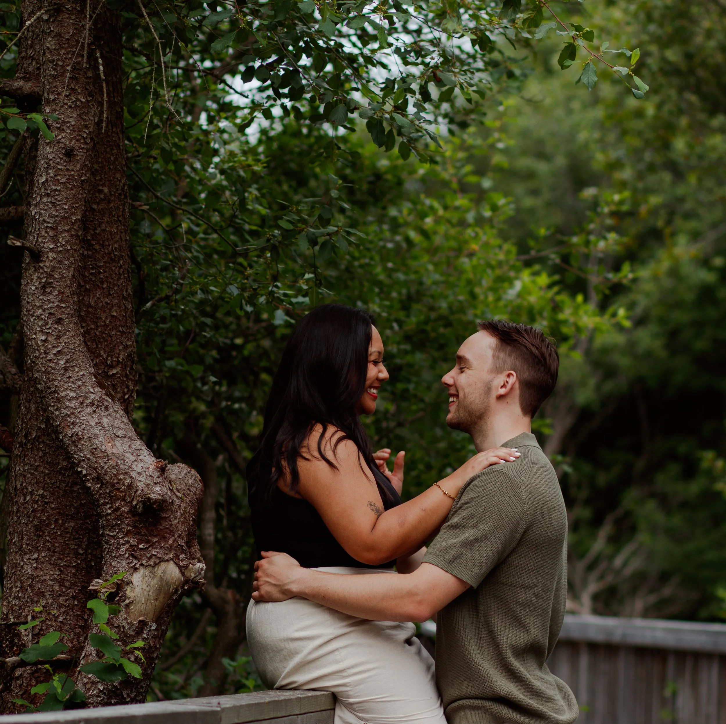 A couple sitting on a wooden railing outdoors, smiling and looking at each other, surrounded by green trees and foliage.