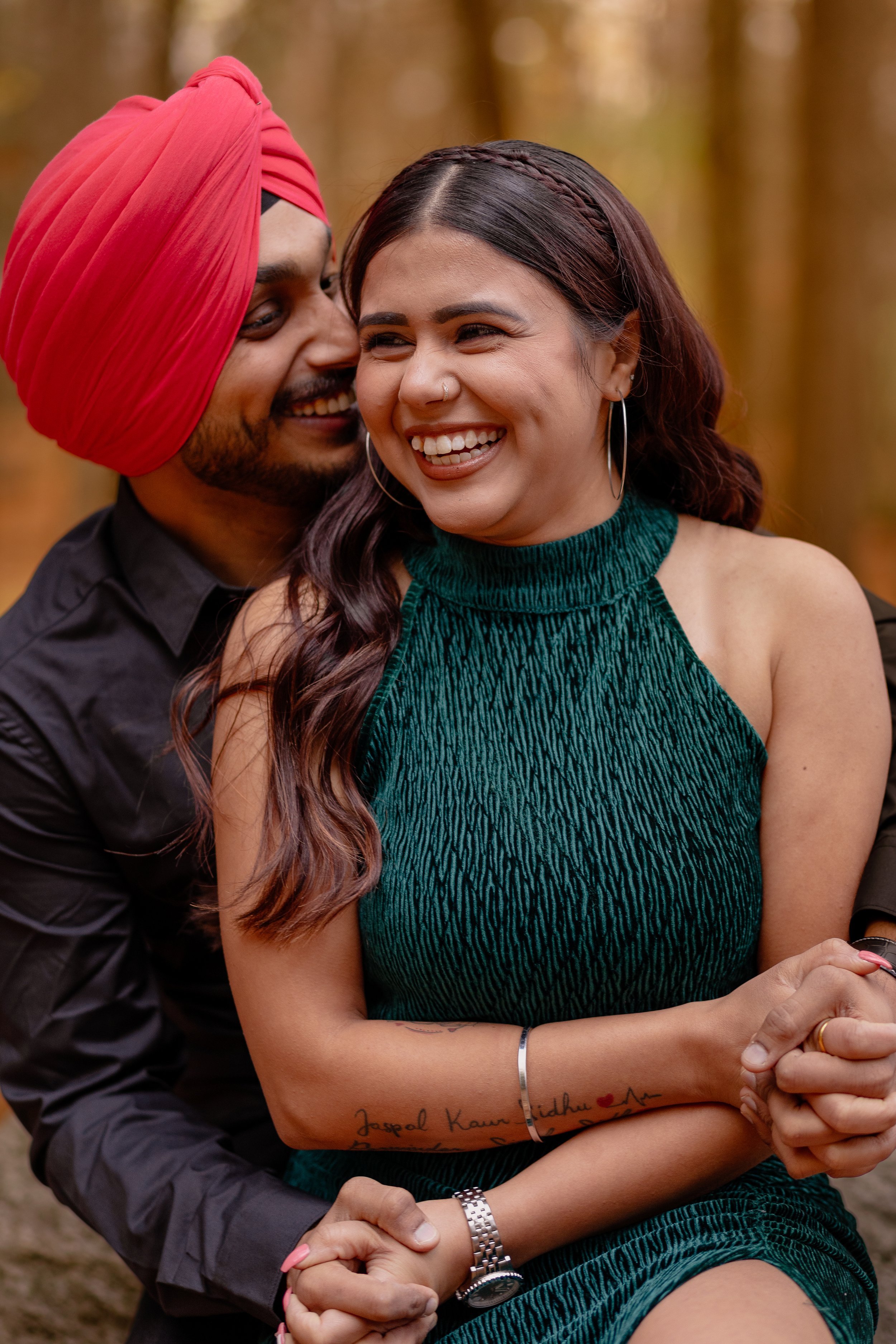 A couple, a man wearing a red turban and a woman wearing a green sleeveless top, are smiling and sharing a moment together outdoors in a forested area.
