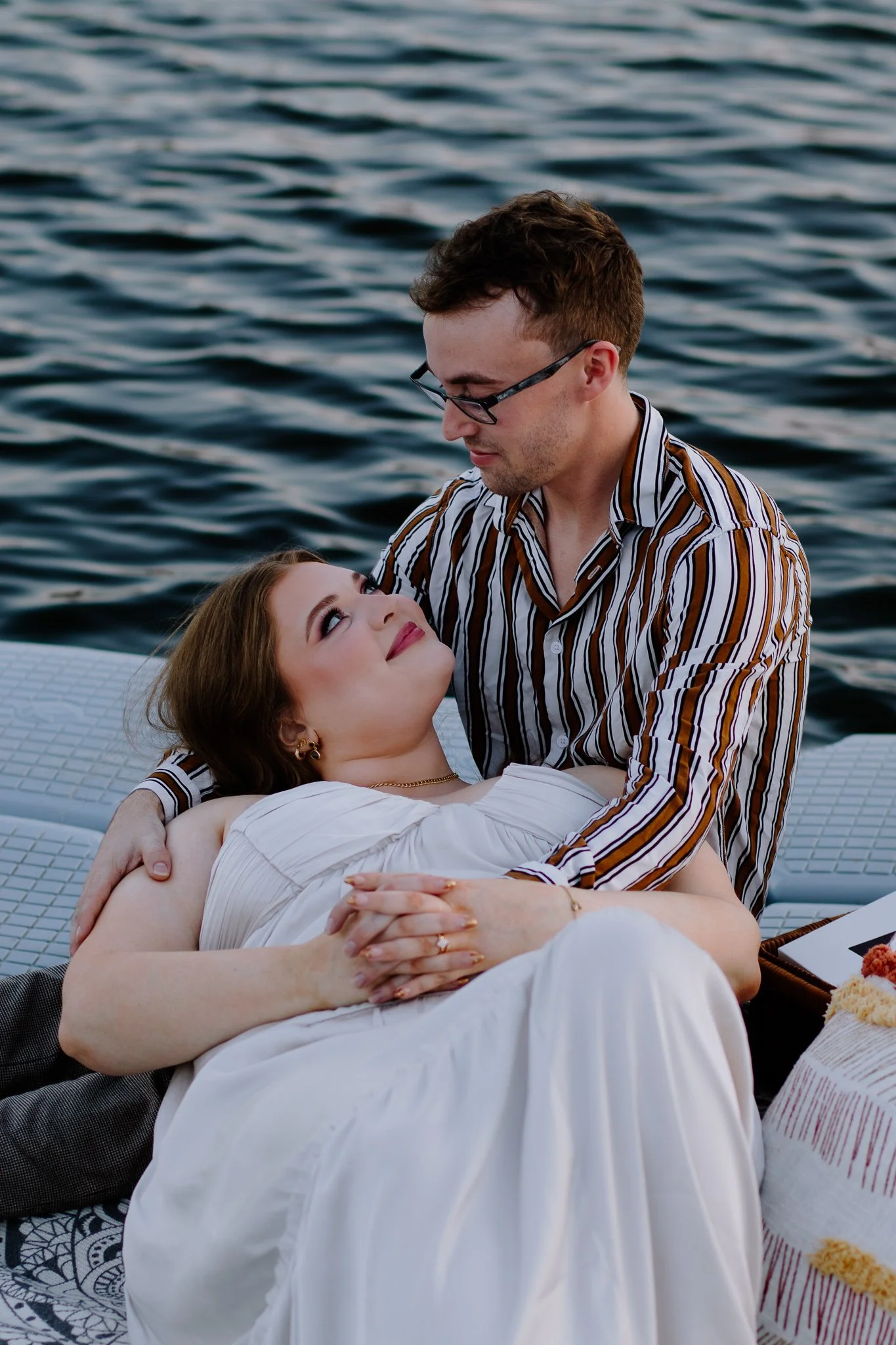A couple sitting by the water, with the woman lying on the man's lap, looking up at him, on a boat or a dock, during sunset.