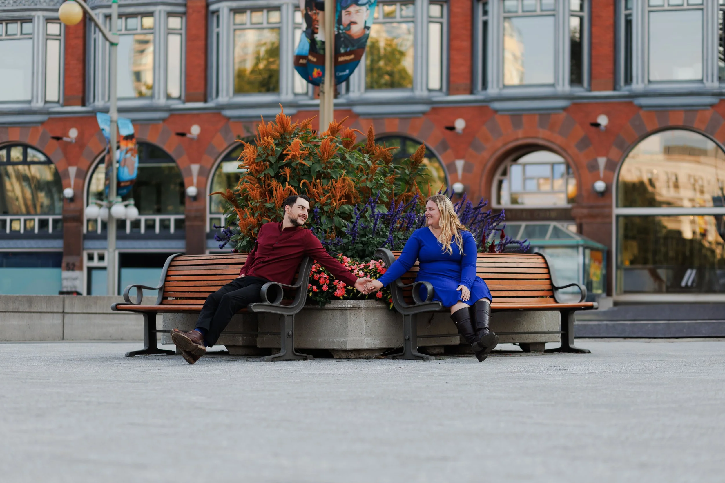 A man and woman sitting on separate benches with a flower planter between them, holding hands and smiling at each other in an outdoor urban setting.