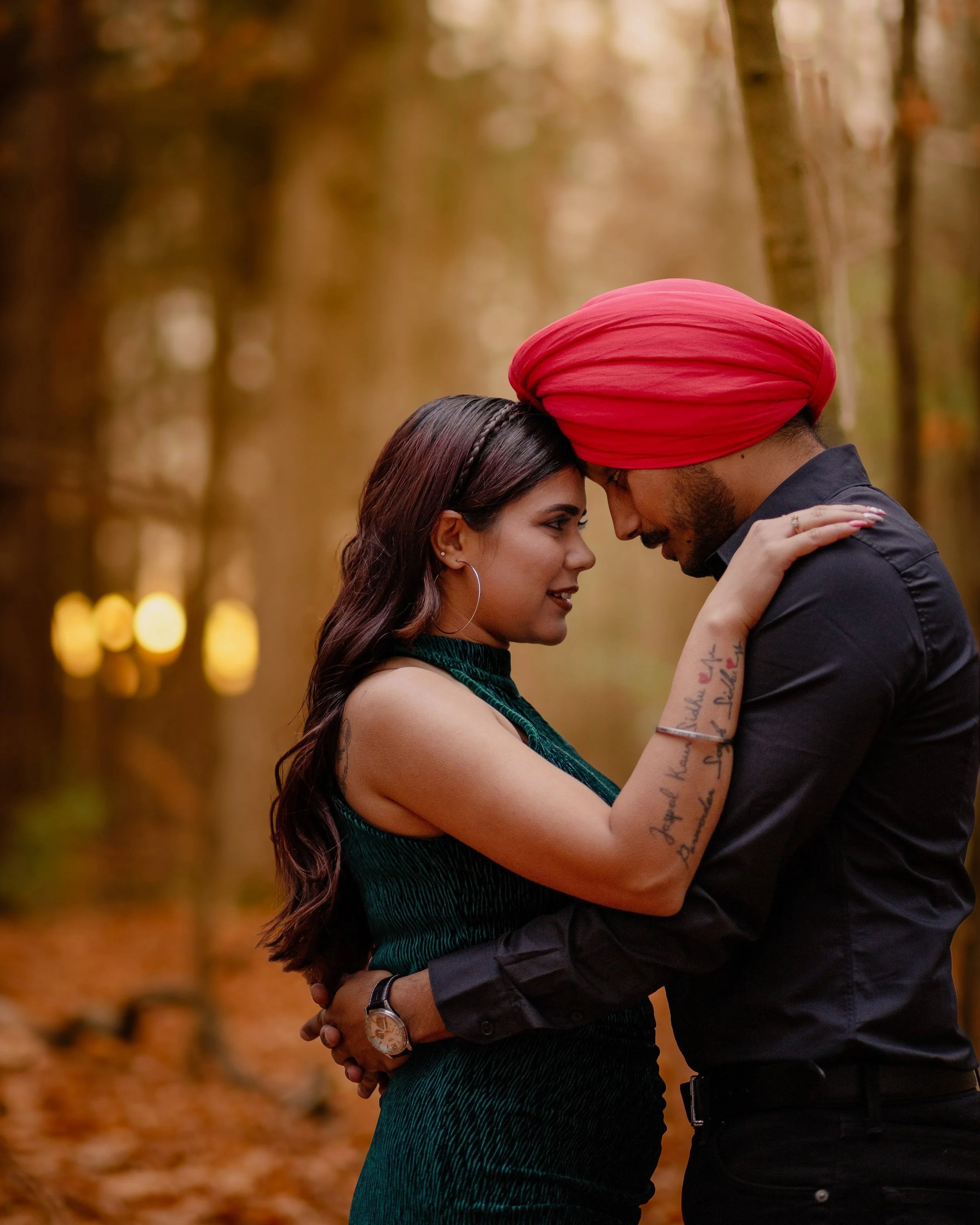 A couple, a woman with long dark hair wearing a green dress and a man with a red turban and black shirt, are close together in an outdoor forest setting with blurred warm-colored trees in the background.