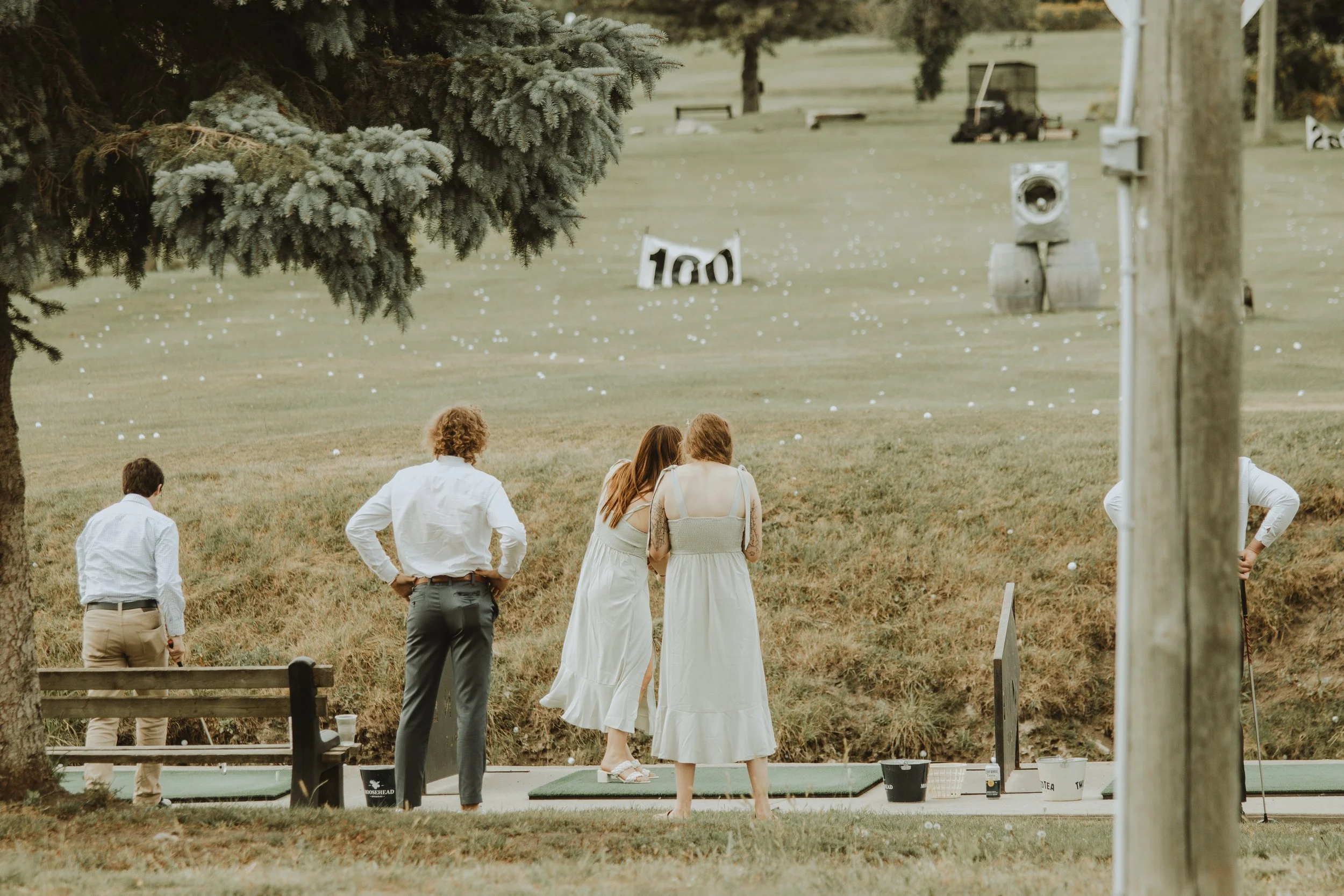 People walking on a putting green at a golf course during daytime, with a large tree in the foreground and a field in the background.