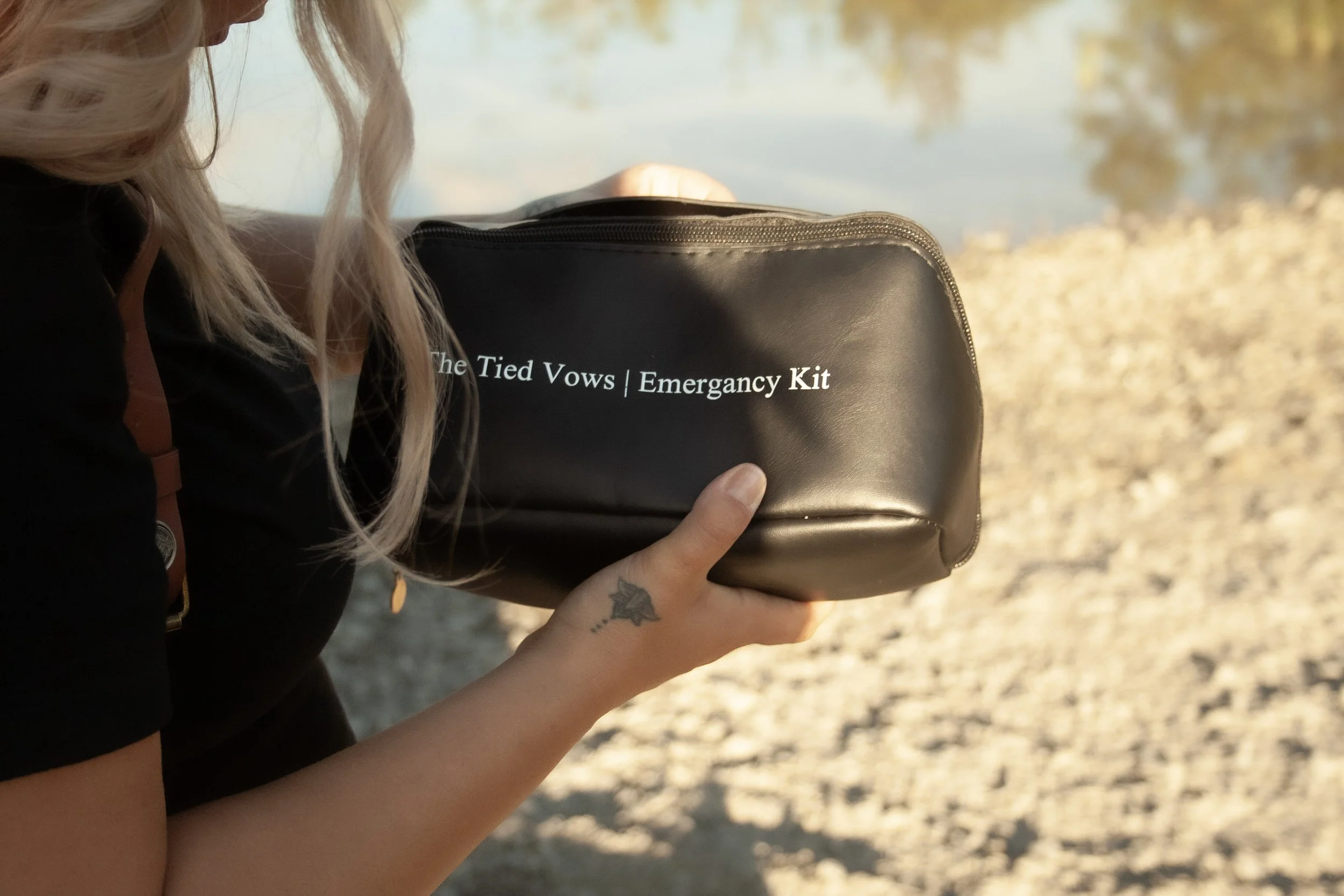 A person with long blonde hair holding a black emergency kit labeled 'The Tied Vows | Emergency Kit' near a body of water in the background.
