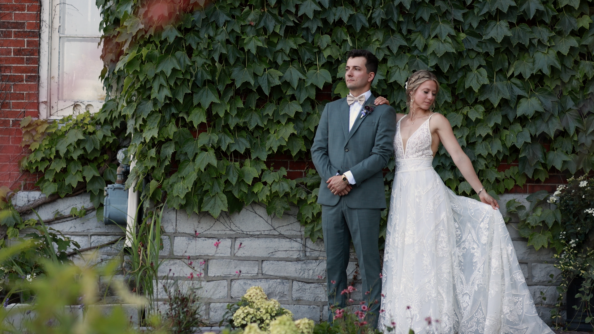 A bride in a white wedding dress with lace details and a groom in a gray suit with a bowtie, standing in front of a brick and ivy wall.