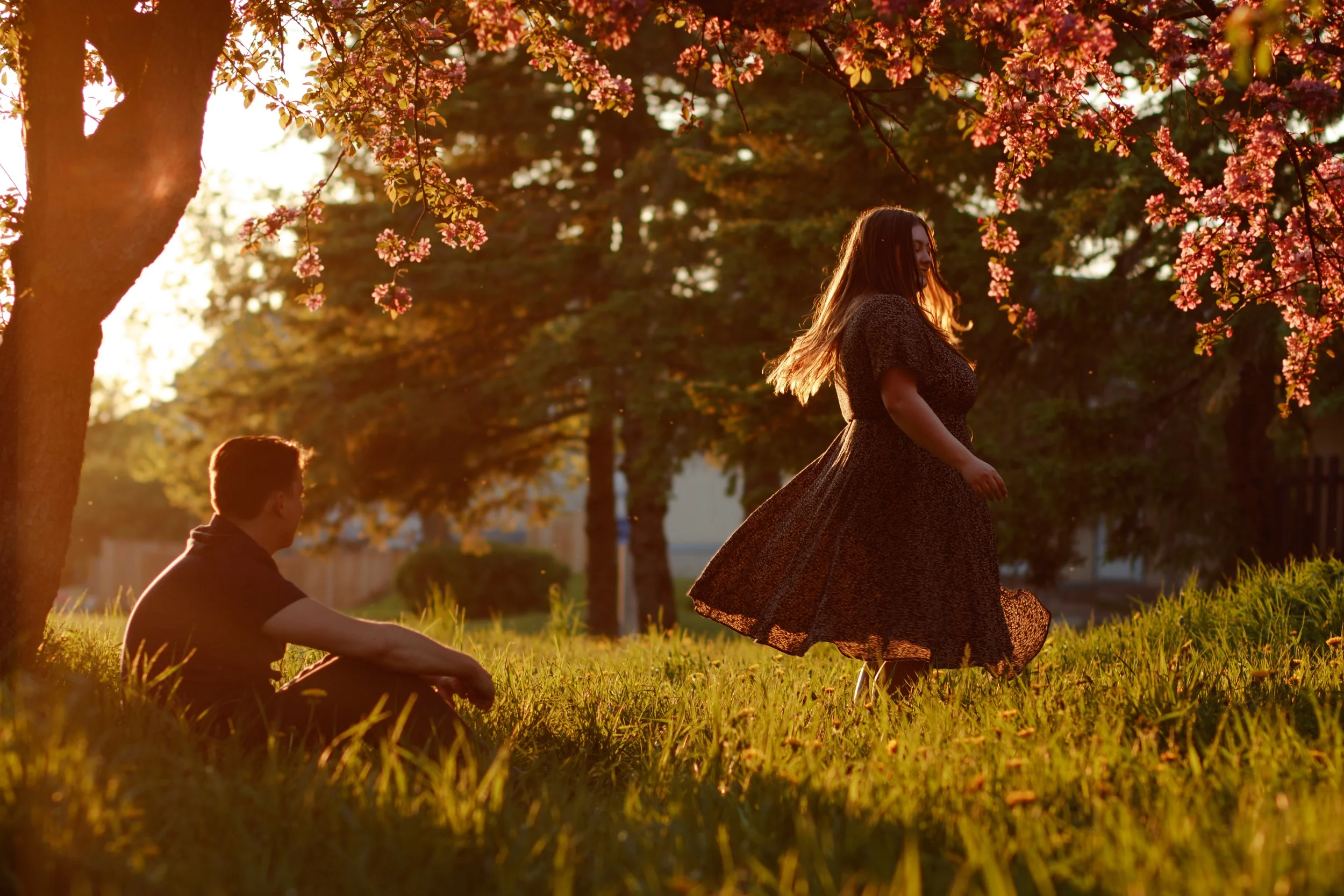 A man sitting on the grass and a woman dancing under cherry blossom trees during sunset.