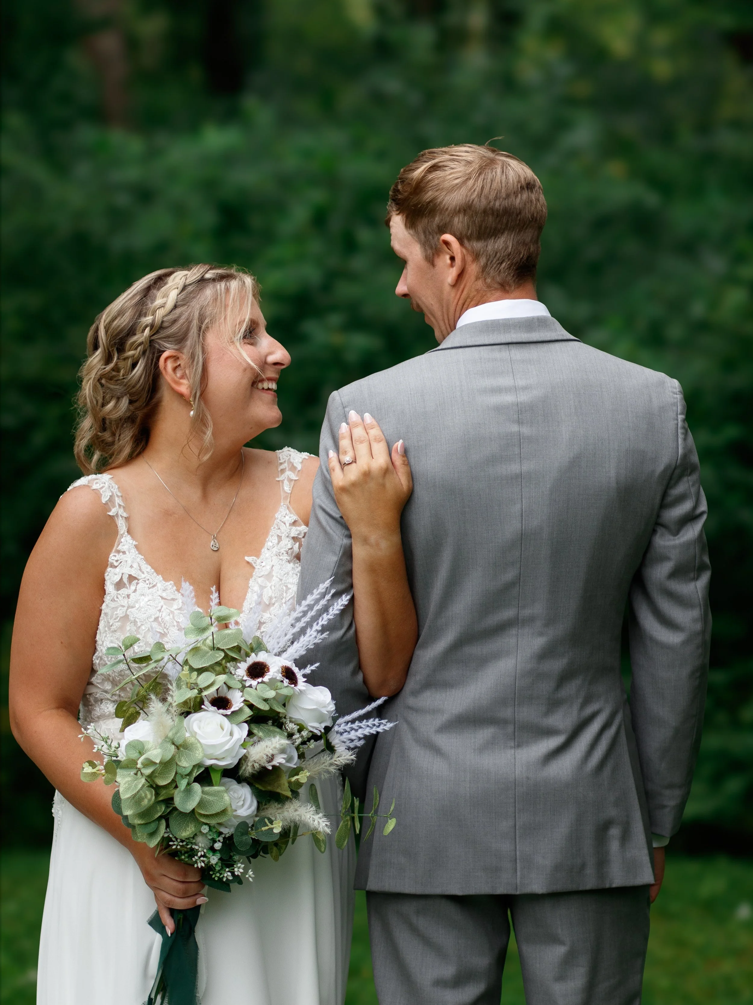 A bride in a white wedding dress holding a bouquet of white flowers and greenery, smiling at a groom in a grey suit, outdoors with green foliage in the background.