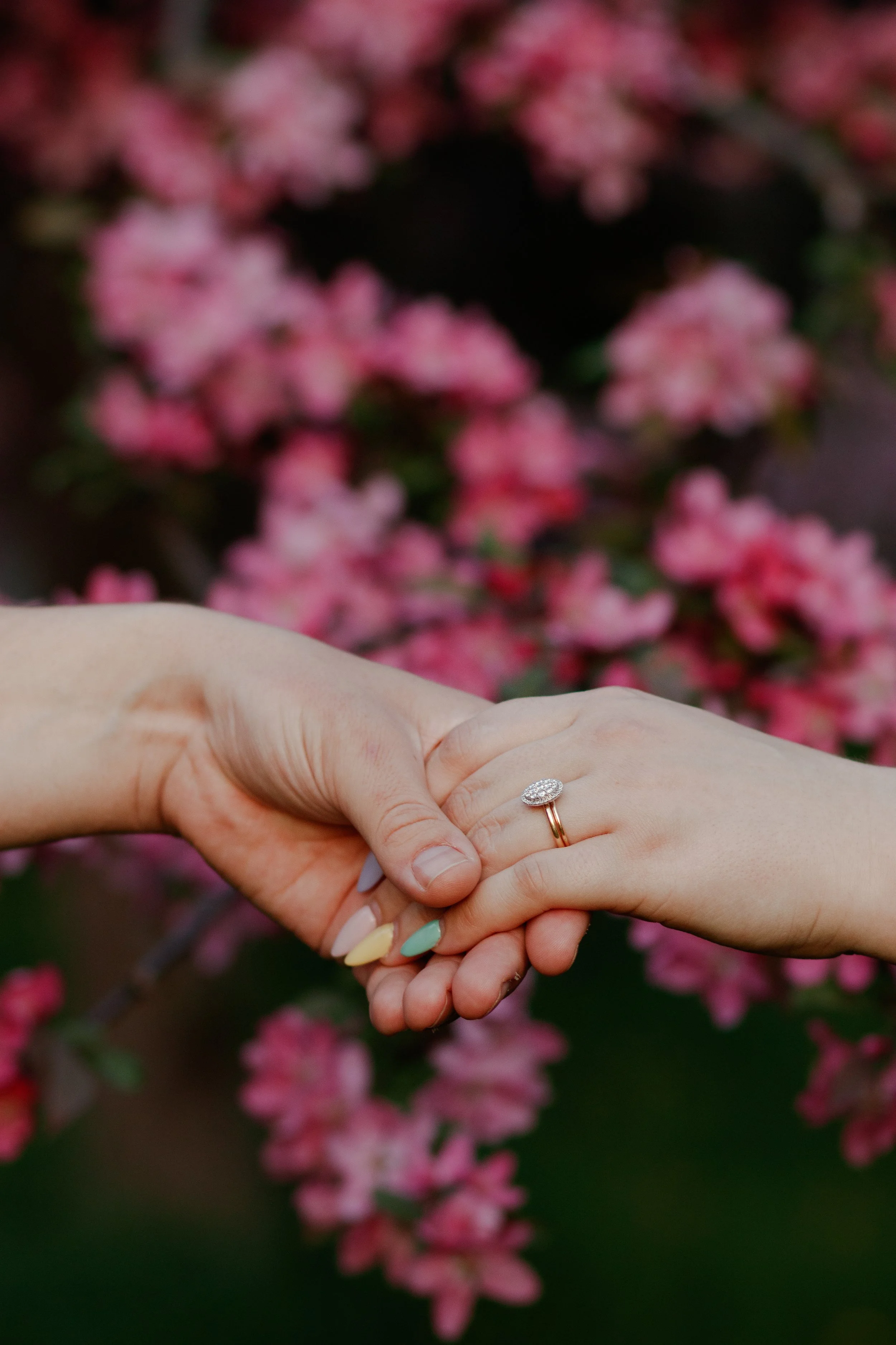 Two hands gently holding each other against a backdrop of pink flowers, with one hand wearing a ring with a large oval-shaped gemstone surrounded by smaller stones.