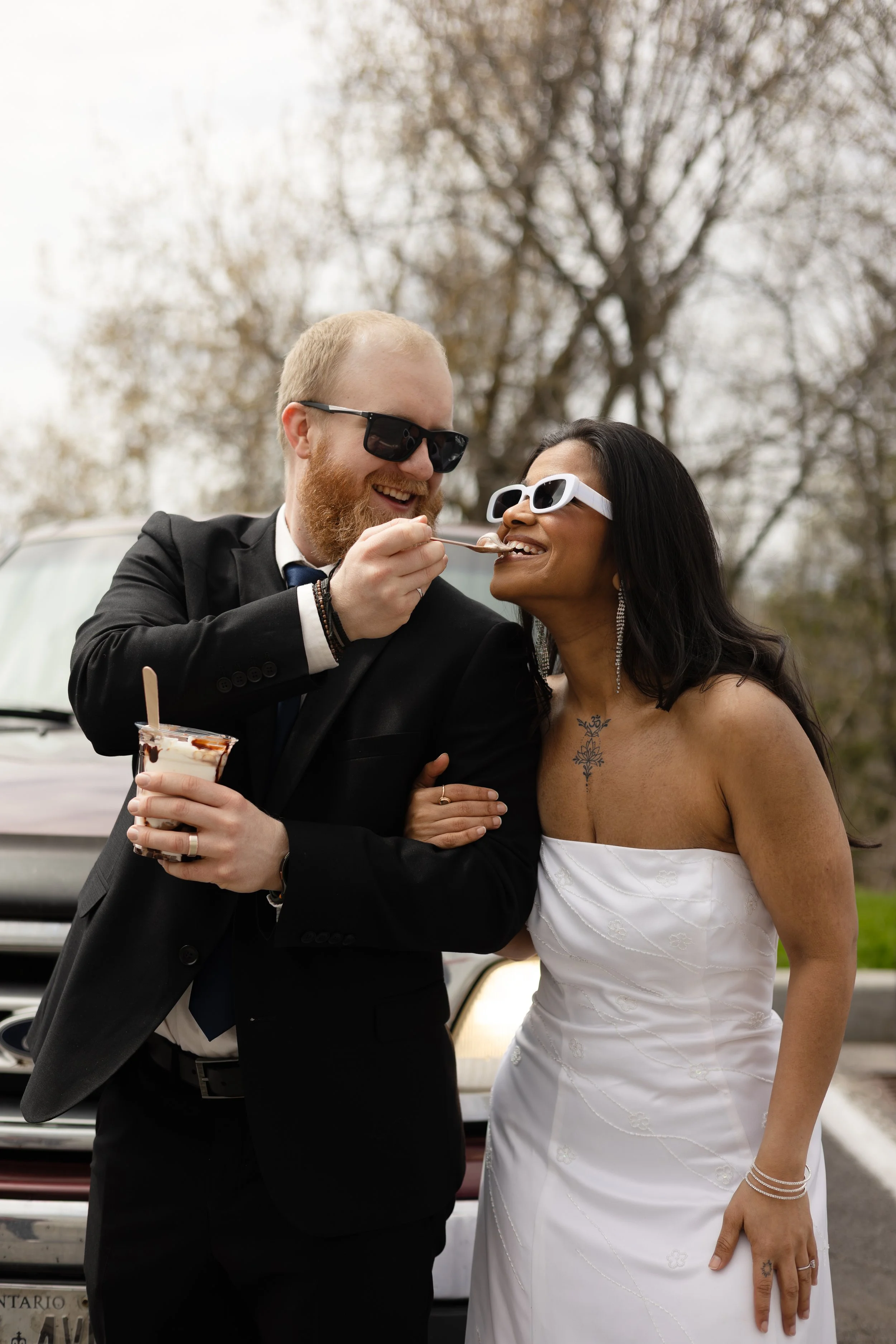 A man in a black suit and sunglasses is feeding a woman in a white strapless dress with a spoon, both smiling and wearing sunglasses, outdoors with a tree in the background.
