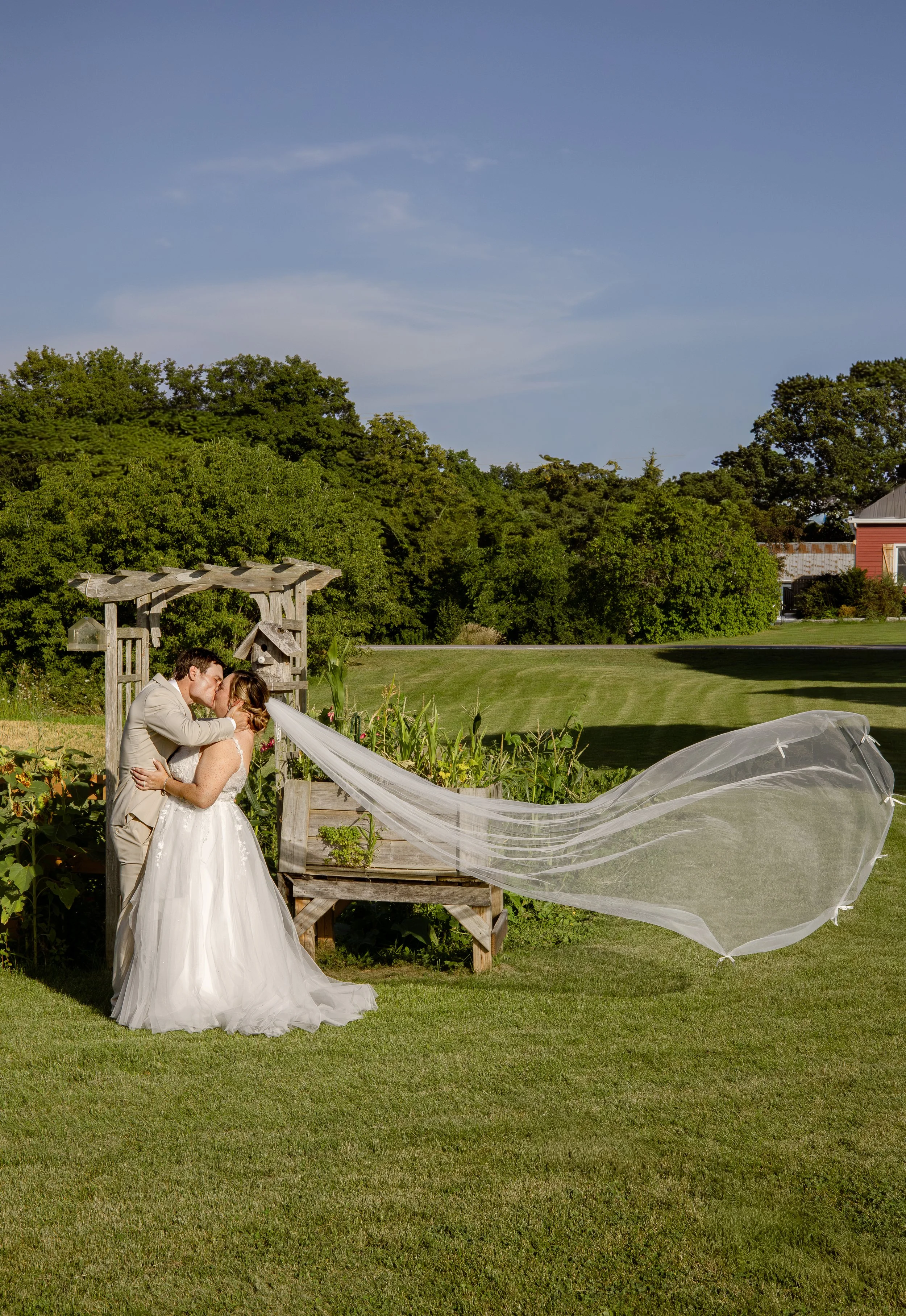 Couple kissing outdoors, bride in a wedding gown and veil, groom in a light suit, near a garden with greenery and a wooden arch.