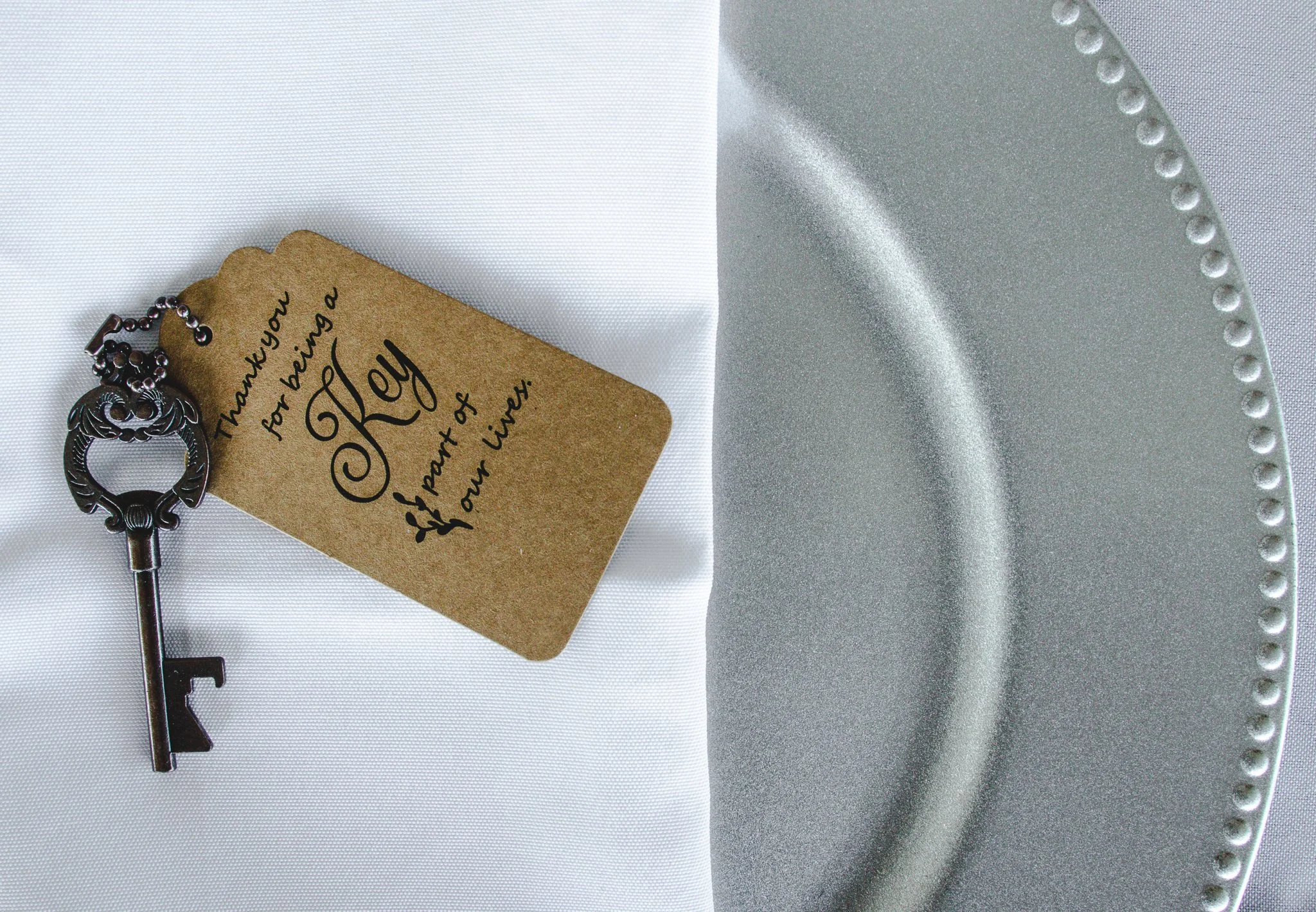 A thank you card that says 'Thank you for being a part of my life, Kelly' next to a vintage-style silver key and a round silver tray with a beaded edge, on a white tablecloth.