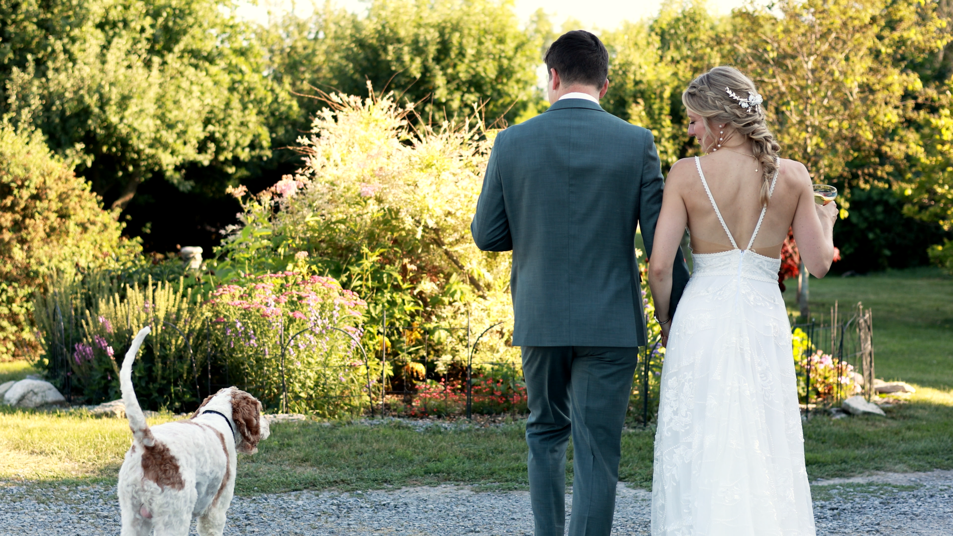 A bride and groom walking hand in hand outdoors, with the bride holding a drink, surrounded by greenery and flowers, with a dog nearby.