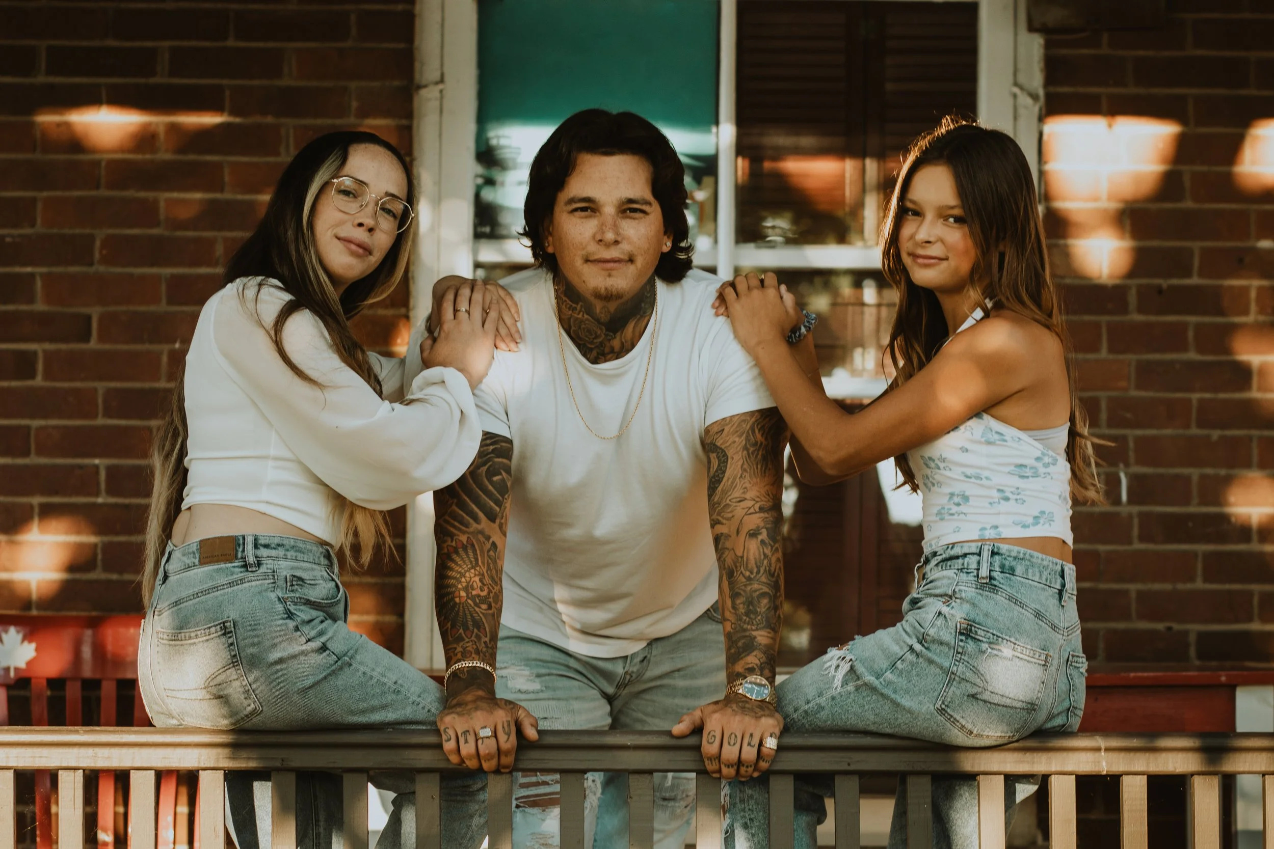 Three young adults in casual clothing posing together in front of a brick wall, with two women kneeling on a wooden railing and a man leaning forward on the railing.