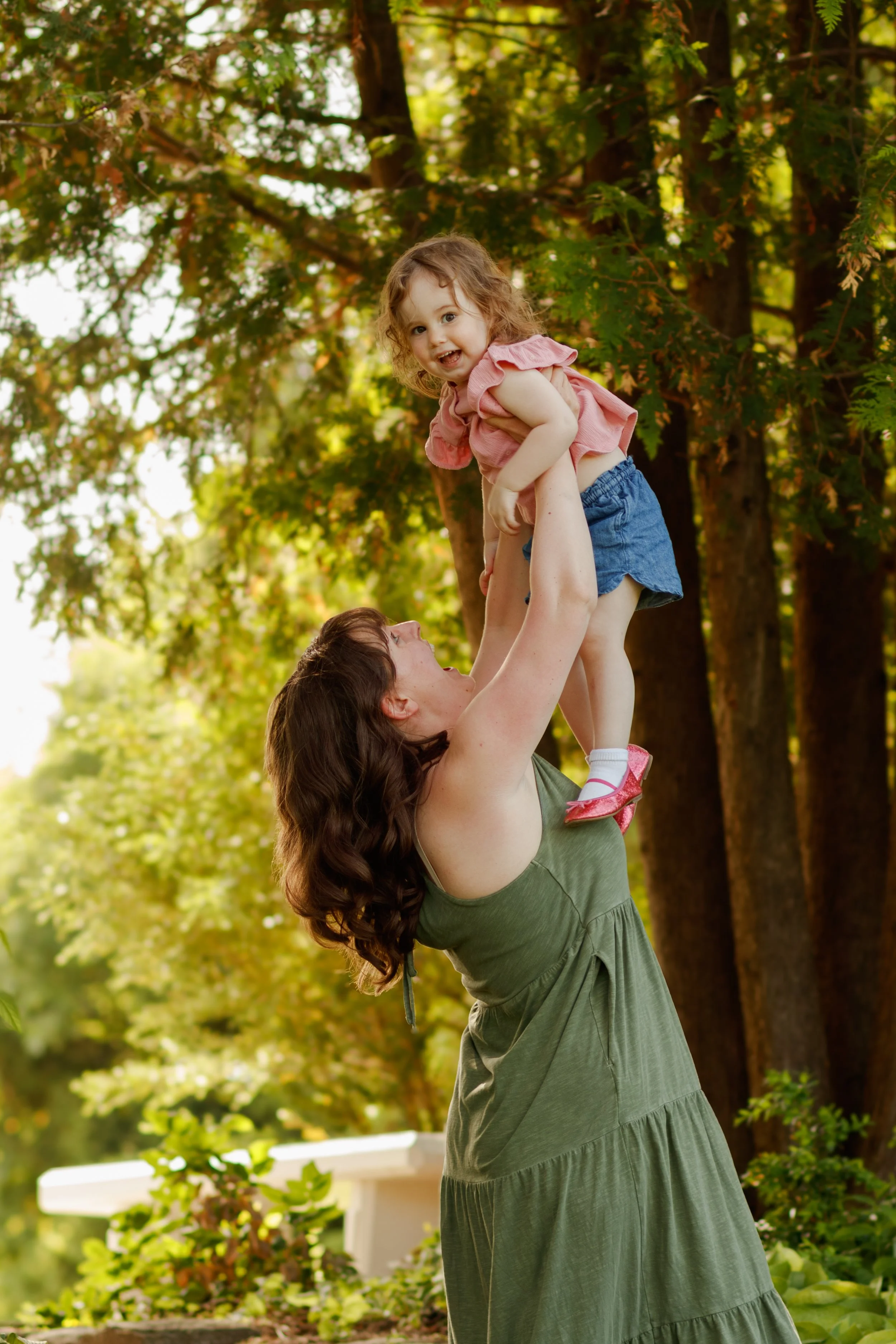 A woman lifting a young girl into the air outdoors, surrounded by trees and greenery.