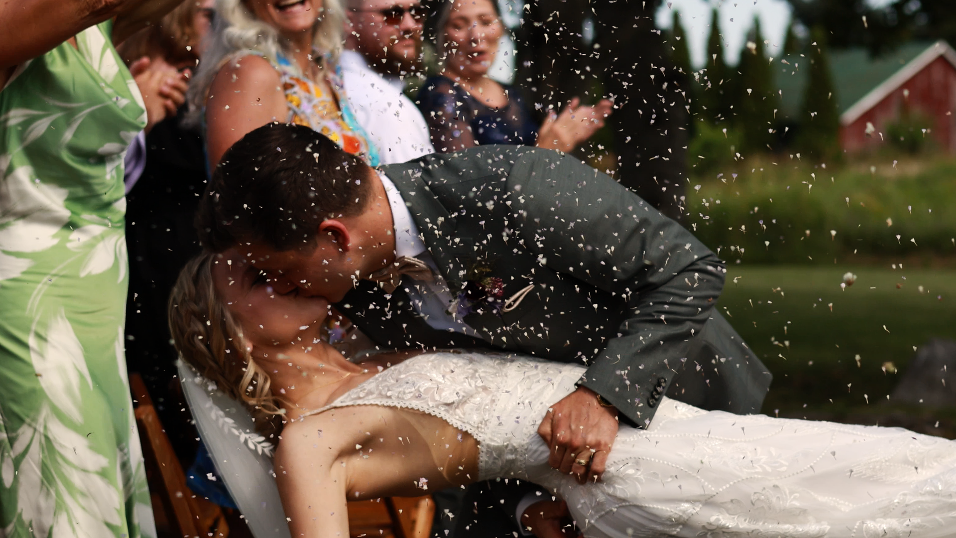 A newlywed couple is kissing during their wedding celebration outdoors, with guests surrounding them and throwing confetti in the air.