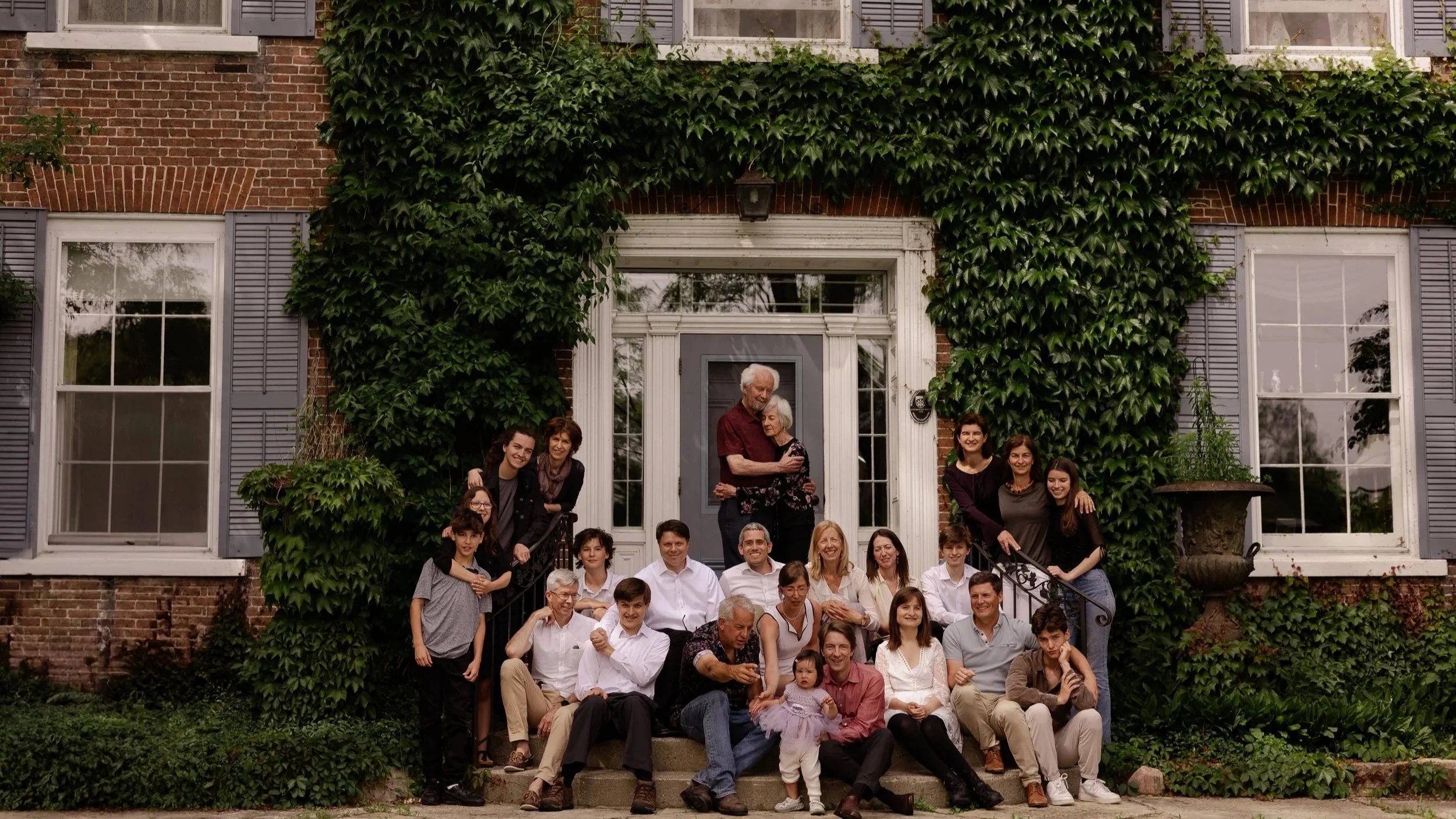 A large family gathering outside a brick house covered with green ivy, with several children, adults, and elderly people smiling and posing for the photo.