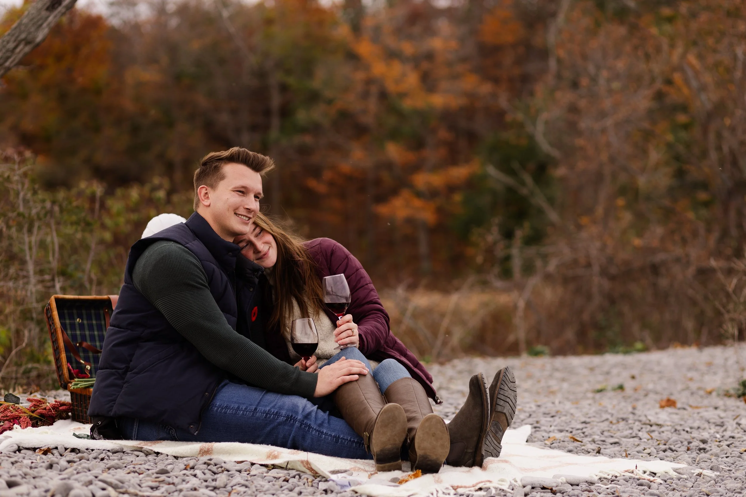 A happy couple sitting on a blanket on a rocky shore, enjoying wine, with autumn trees in the background.