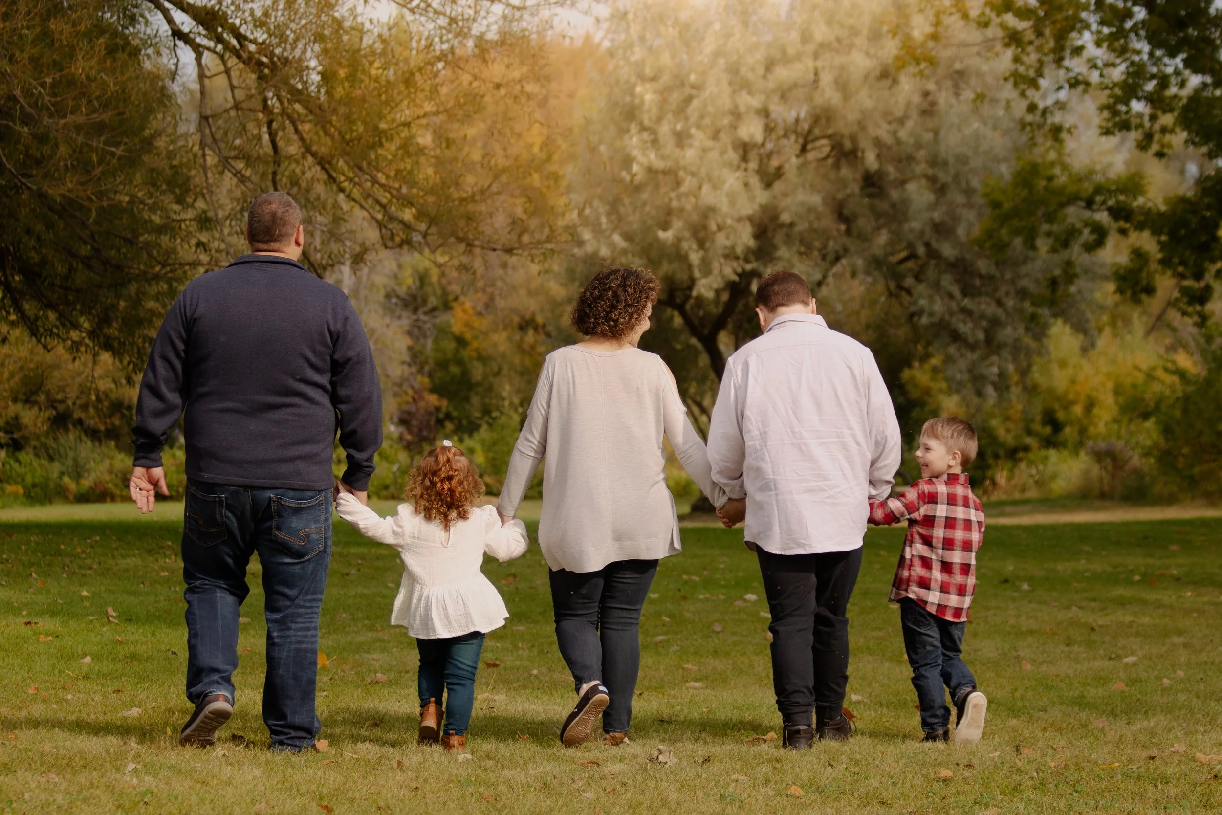 A family walking in a park during autumn, holding hands. The group includes two adults and three children, with colorful fall foliage in the background.