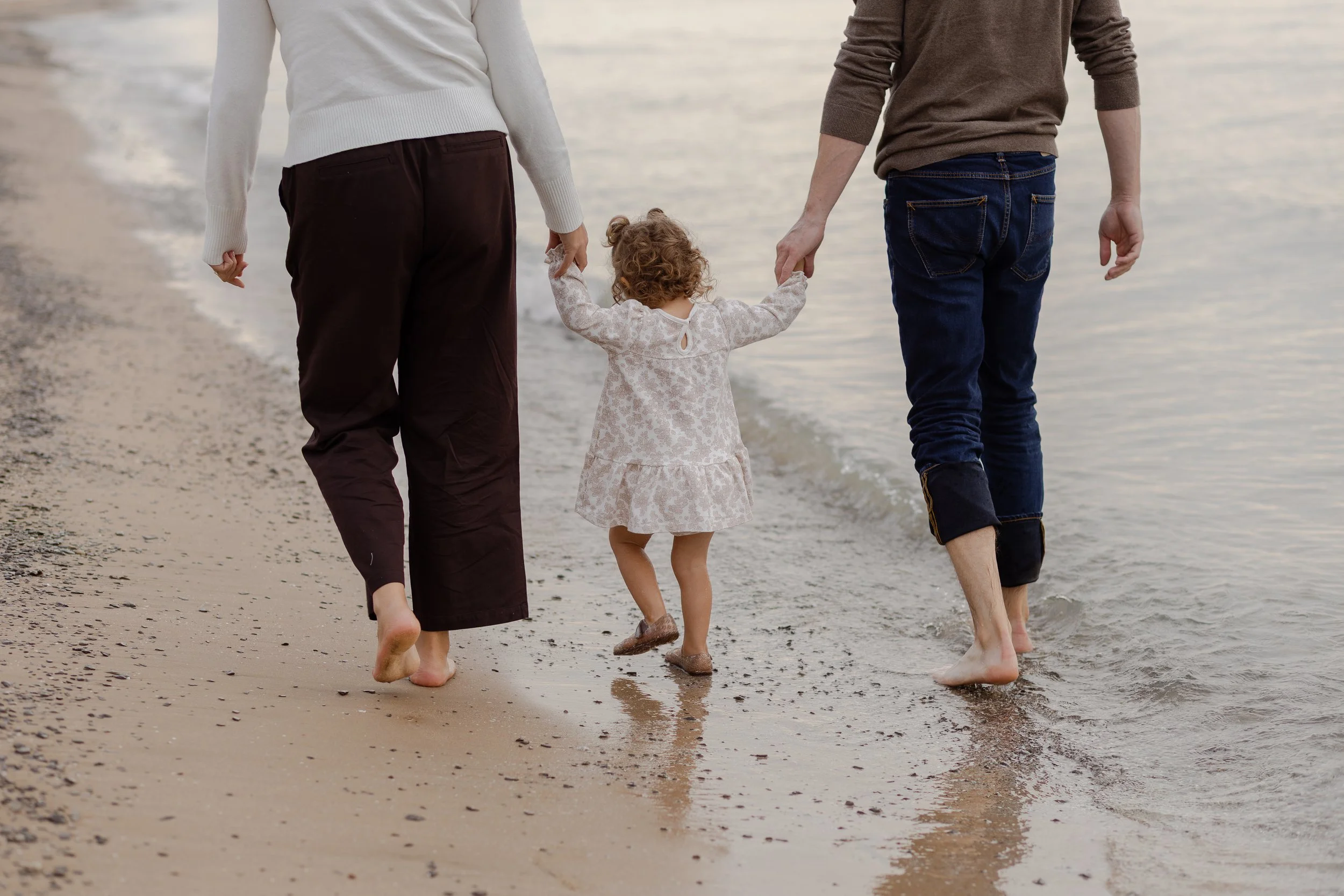 A family of three small children walking barefoot along the shoreline of a beach with waves gently washing up.