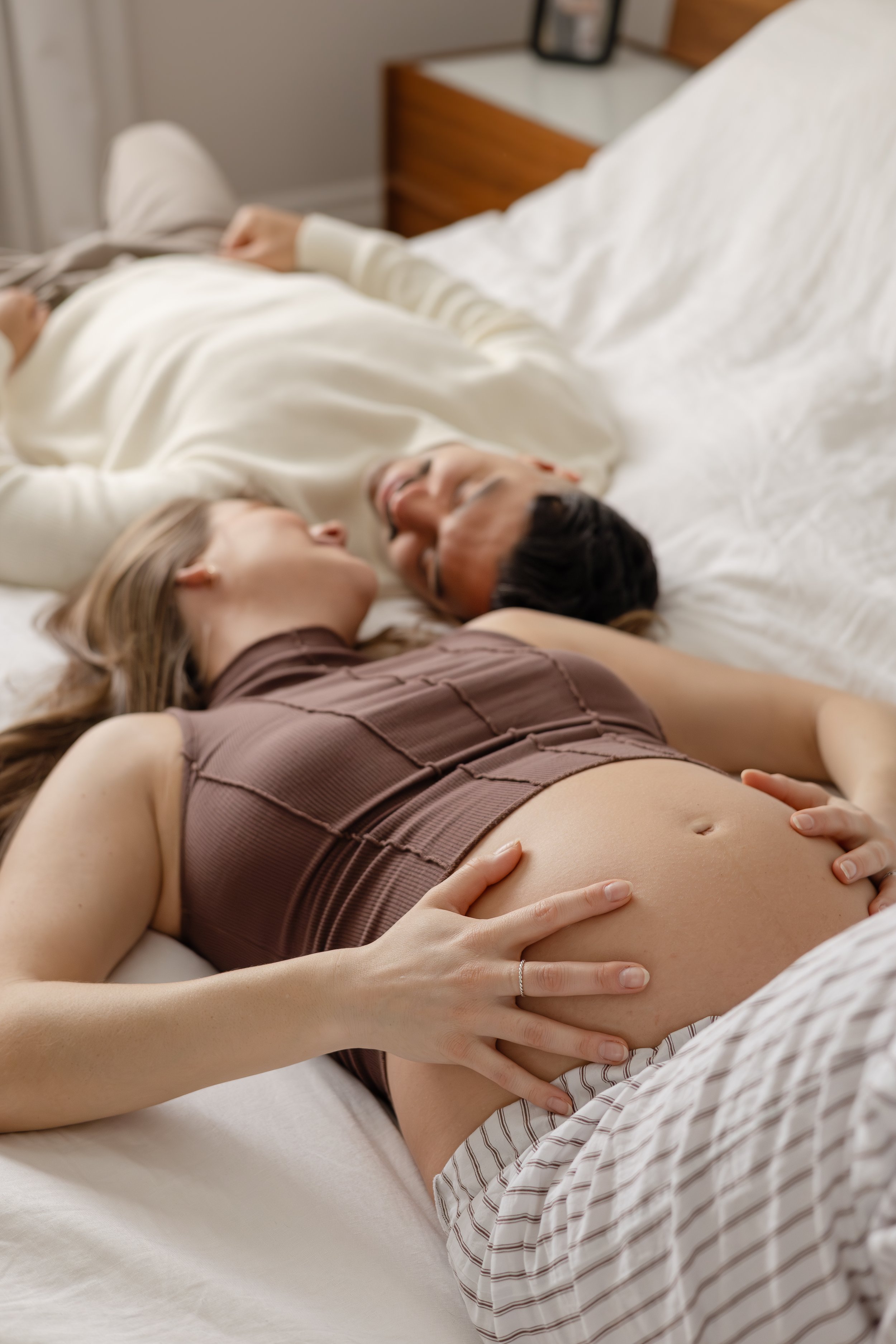 A pregnant woman lies on her back on a bed with her hand resting on her belly, smiling and looking at a woman next to her, both lying close together.  by The Tied Vows Ontario  Wedding Photography, Film & Bridal Assistant Company