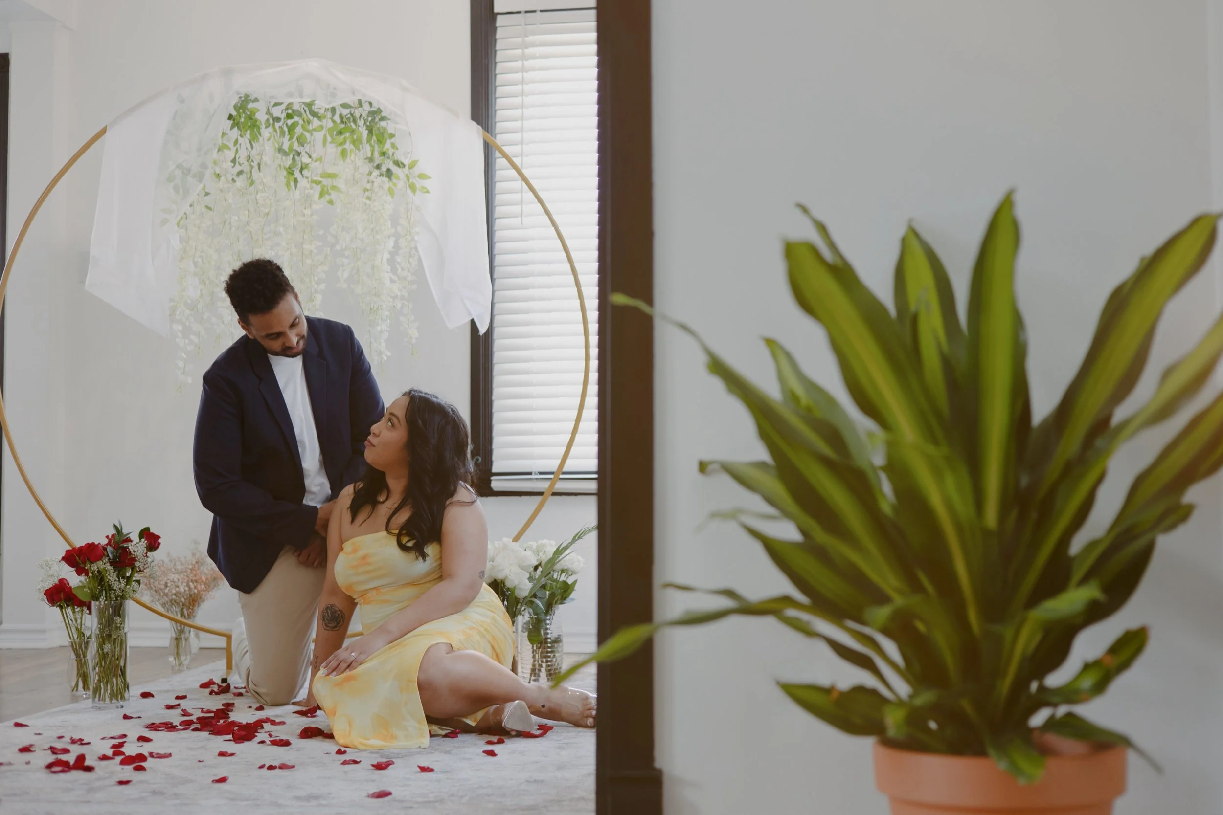 A man kneeling next to a woman sitting on the floor with rose petals scattered around. The setting has flowers and plants, and there's a circular frame with hanging greenery behind them.