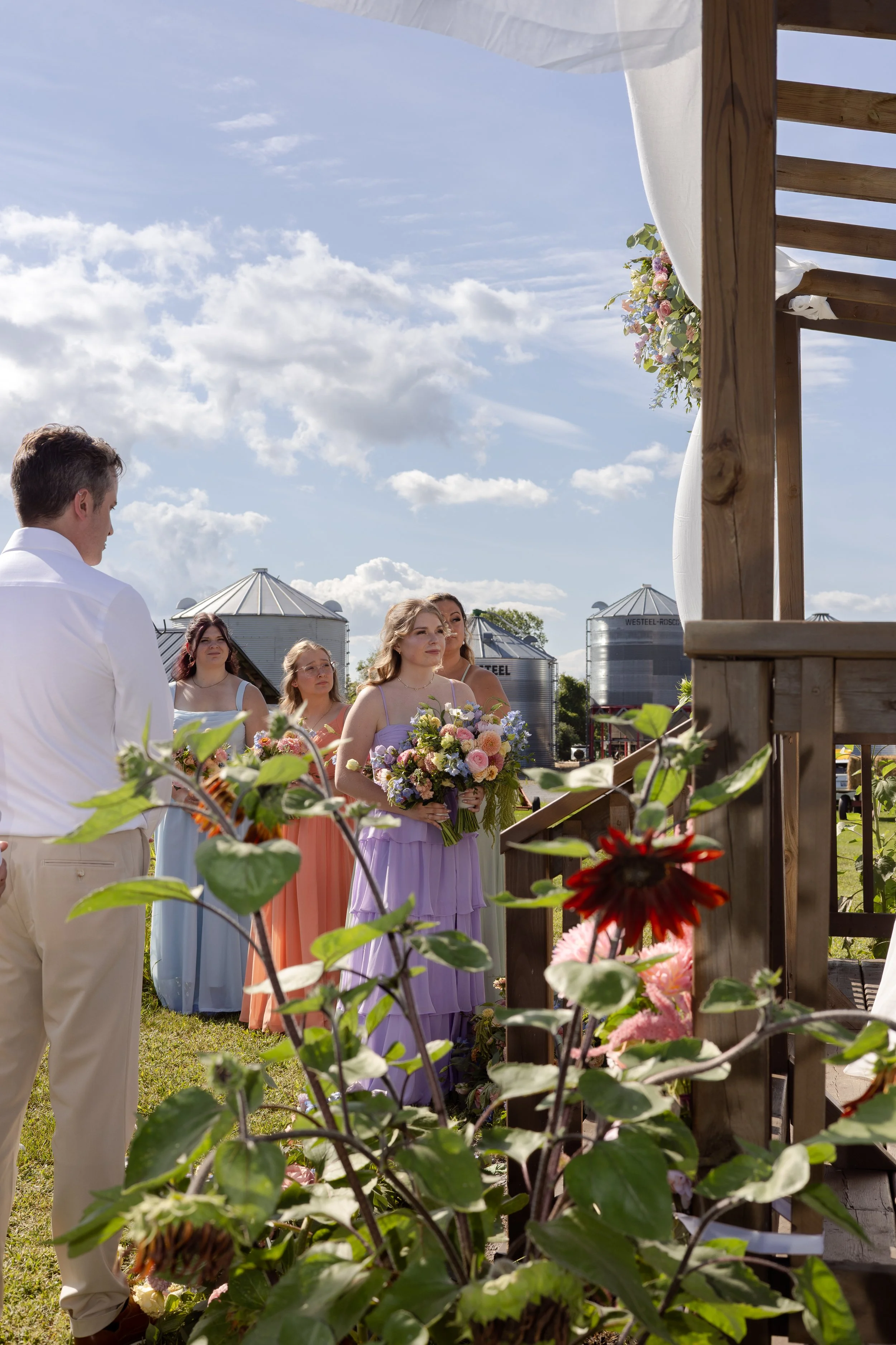 A wedding ceremony outdoors with a group of people, including bridesmaids holding bouquets, under a decorated wooden arch with flowers and fabric. Grain silos and blue sky in the background.