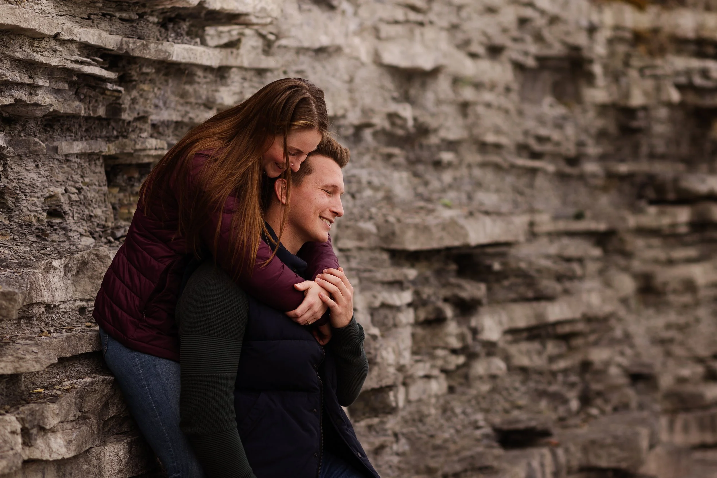 A woman riding on a man's back, both smiling, against a rocky background.