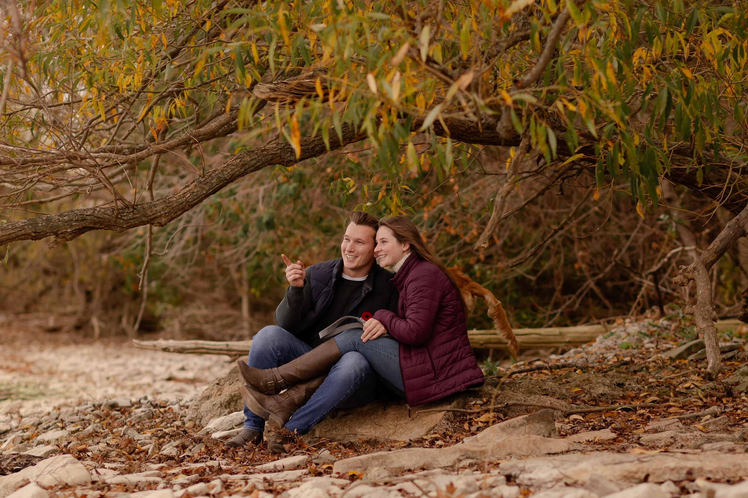 A man and woman sitting on a large rock outdoors, surrounded by autumn foliage, smiling and sharing a happy moment.