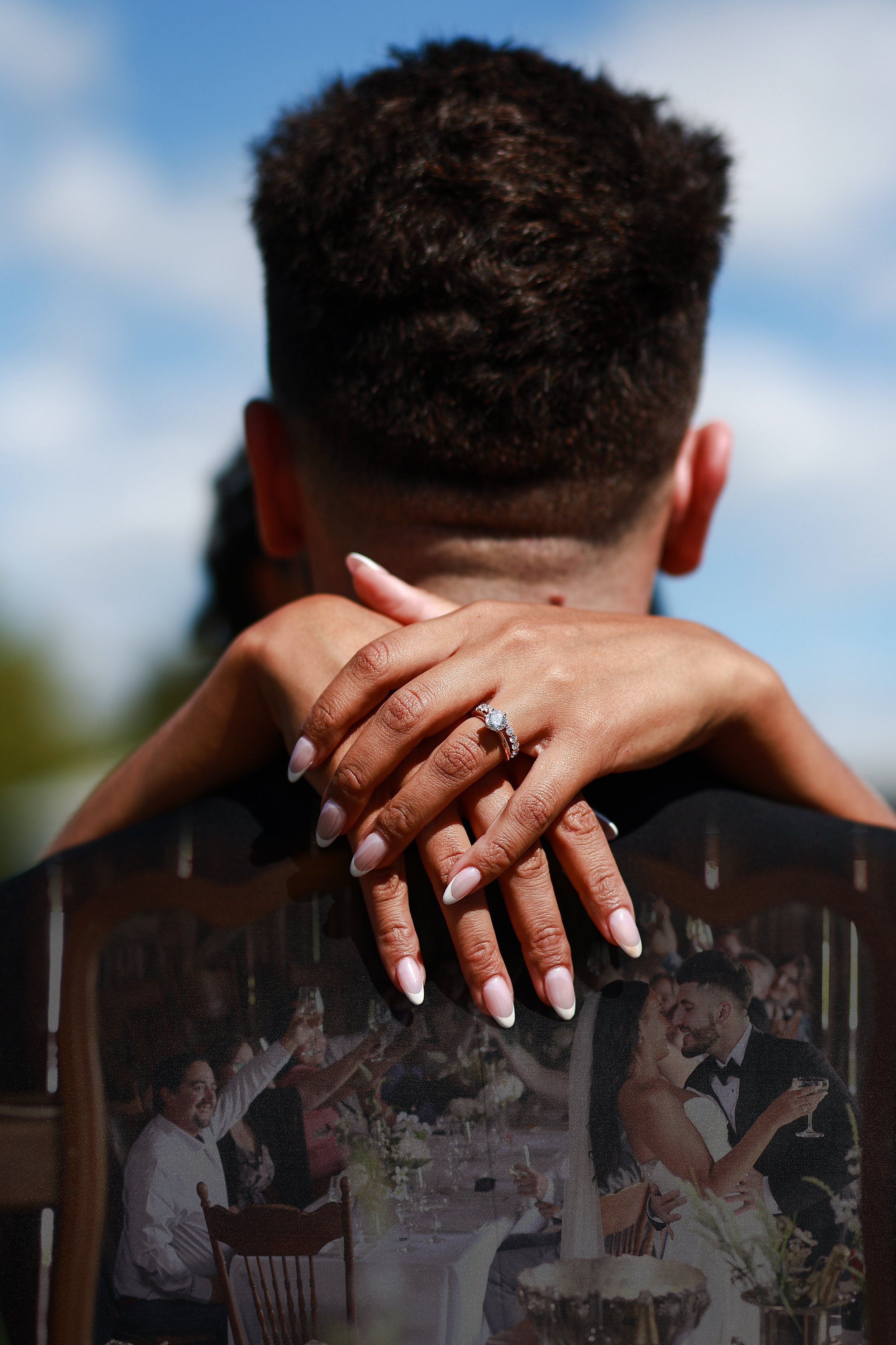 A woman's hands with a wedding ring on her finger are wrapped around a man's neck, with a wedding photo reflected on her nails showing a wedding celebration and a couple dancing.