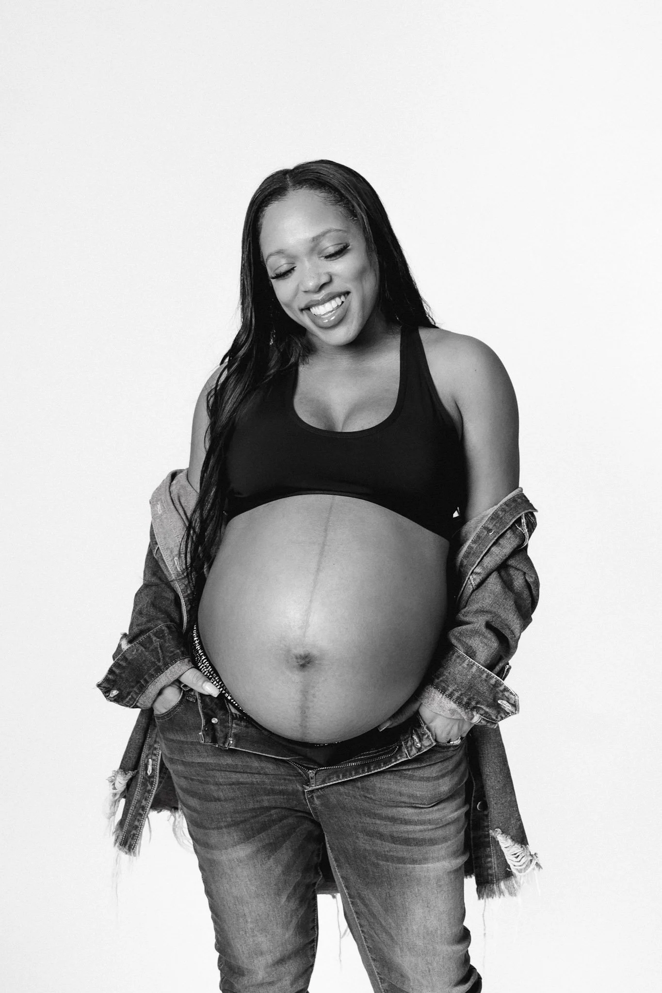 photo of young black pregnant woman wearing denim and black sports bra with denim jacket posing for maternity photoshoot. black and white photo. photo taken by black woman maternity photographer with photo studio in pontiac, mi