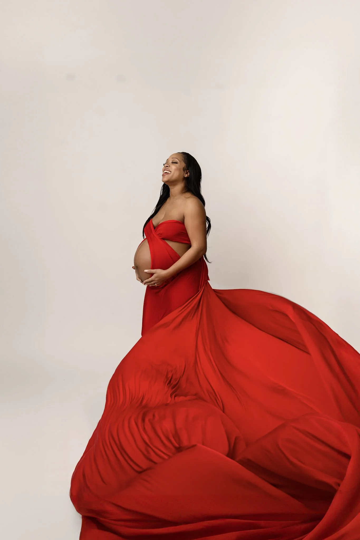 Photo shows young black woman with long black hair wearing a billowing red flying dress posing while holding her pregnant belly. Maternity photoshoot was coordinated by blackw oman potographer in pontiac, michigan who serves the metro detroit area
