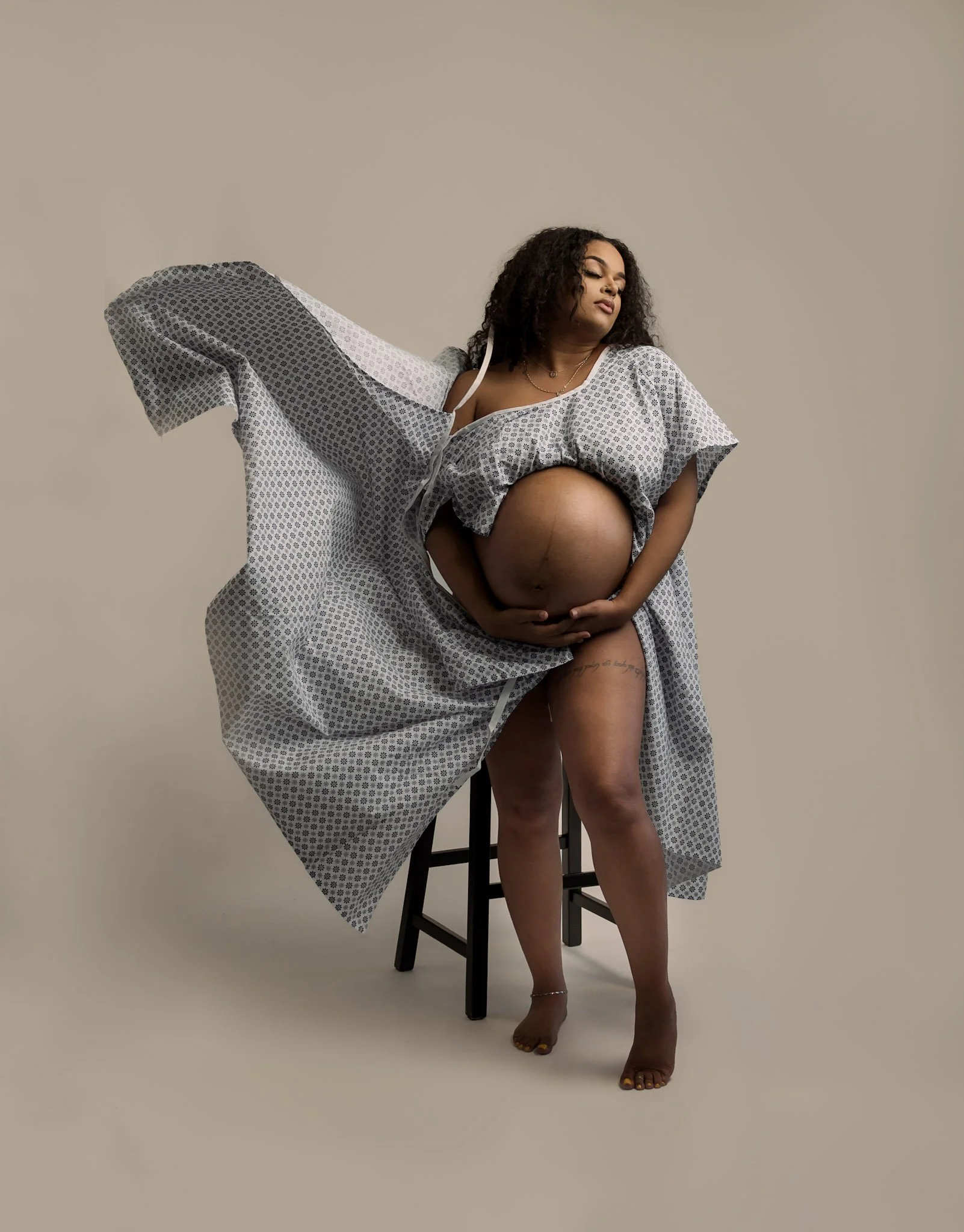 photo of pregant black woman posing for maternity photoshoot wearing a hospital gown. photo was taken by black woman maternity photographer with a photo studio in pontiac, michigan