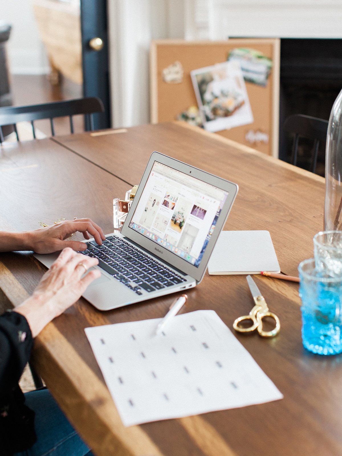 Person working on a laptop at a wooden table with scissors, a notepad, and a glass of water, in a bright room.