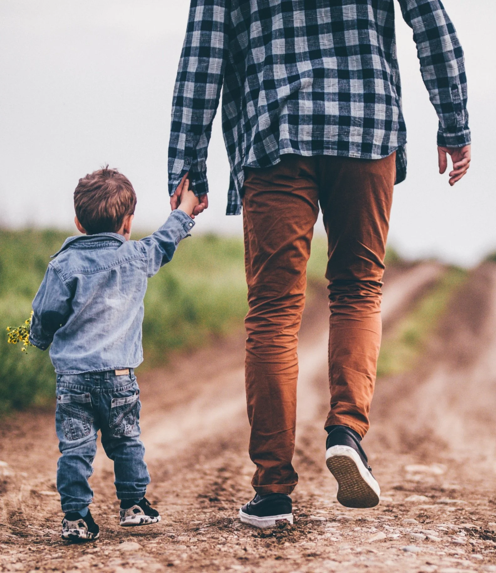 Adult and child walking on dirt path, holding hands, wearing casual clothes.